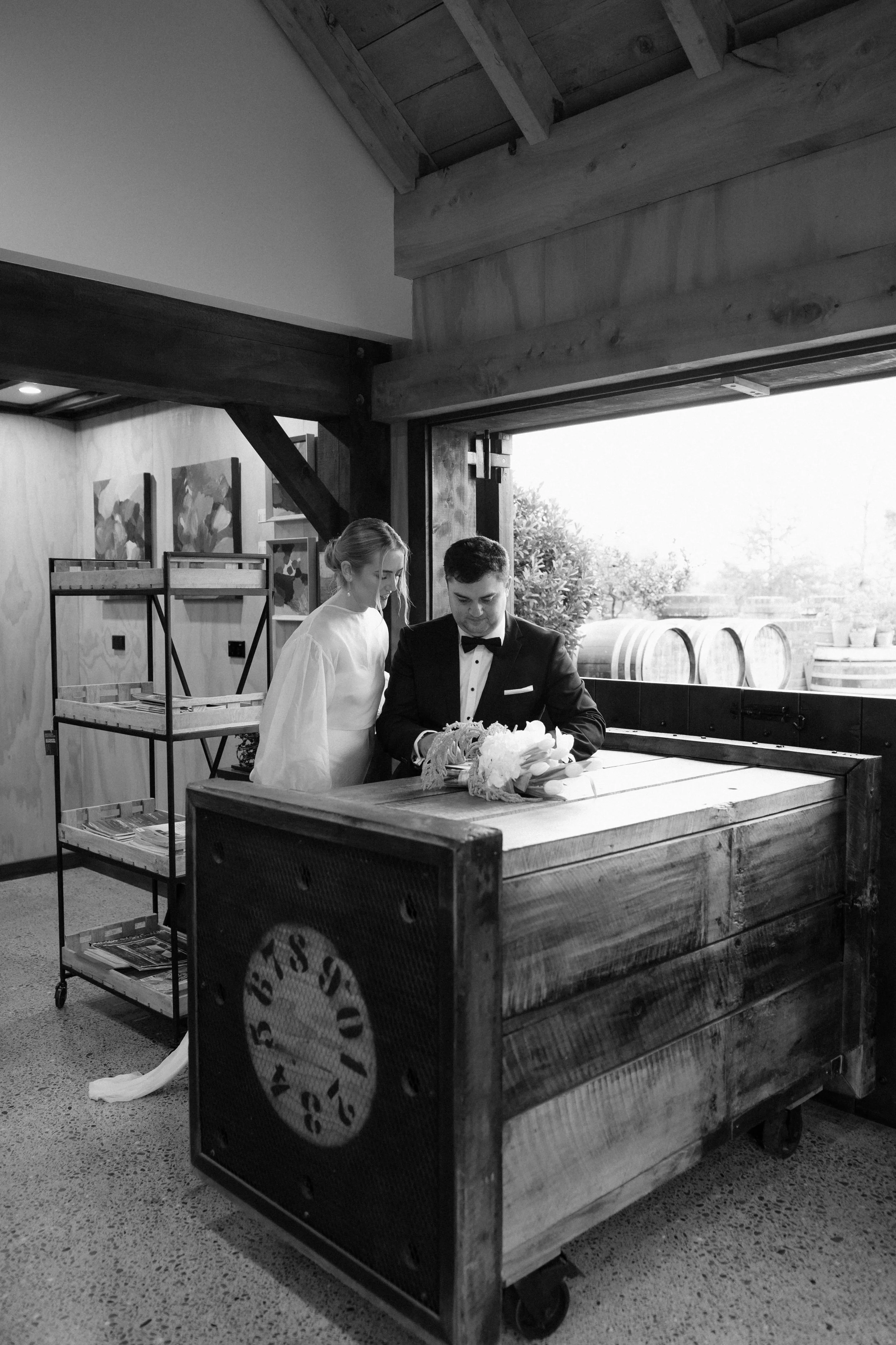A man in a tuxedo and a woman in a dress are standing next to each other, looking down, inside a rustic building with large windows and wooden beams, next to a wooden cart with magazines and a circular logo.