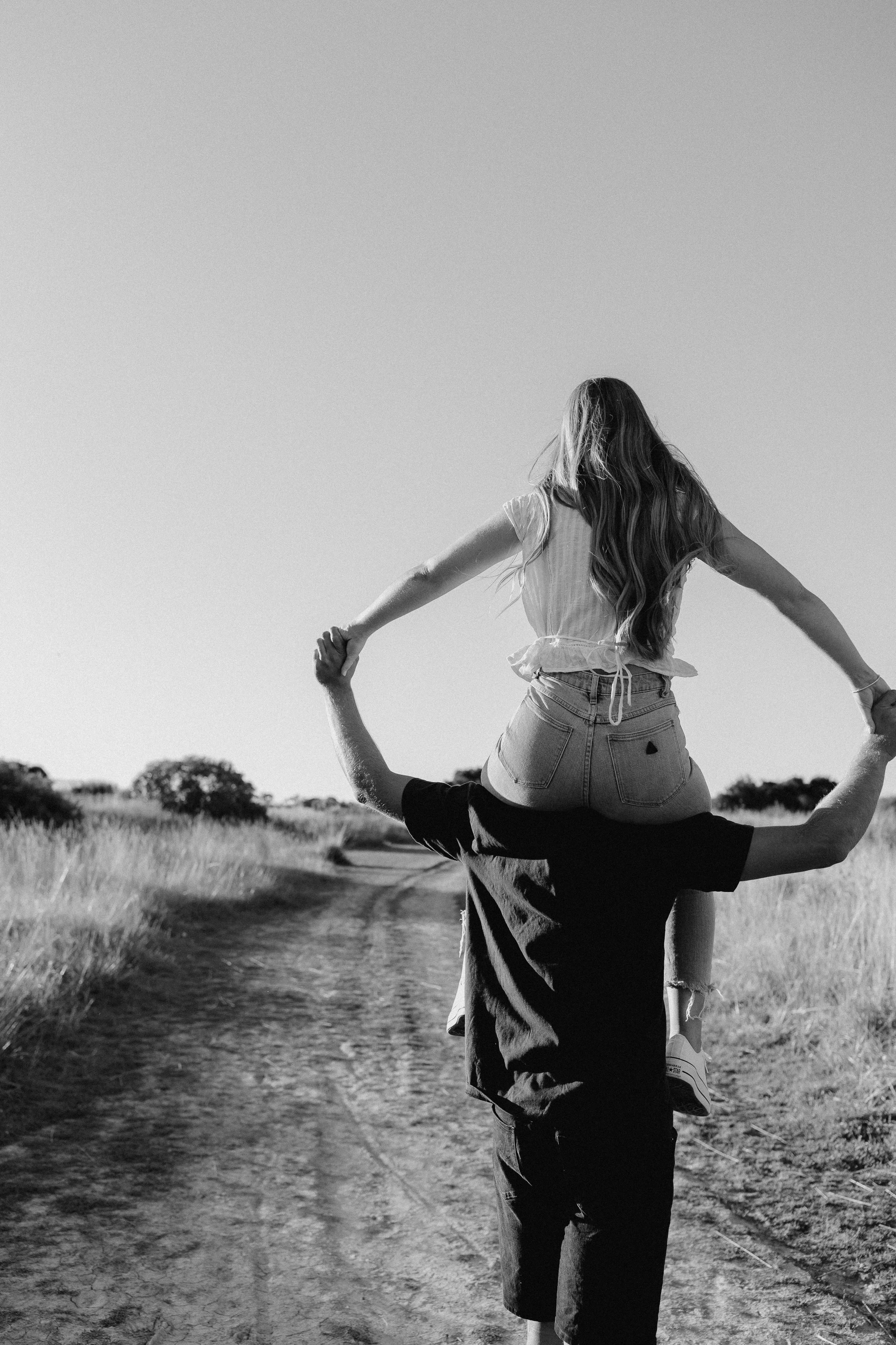 A person standing on the shoulders of another person, holding hands during a yoga pose outdoors on a dirt path with trees in the background, under a clear sky.