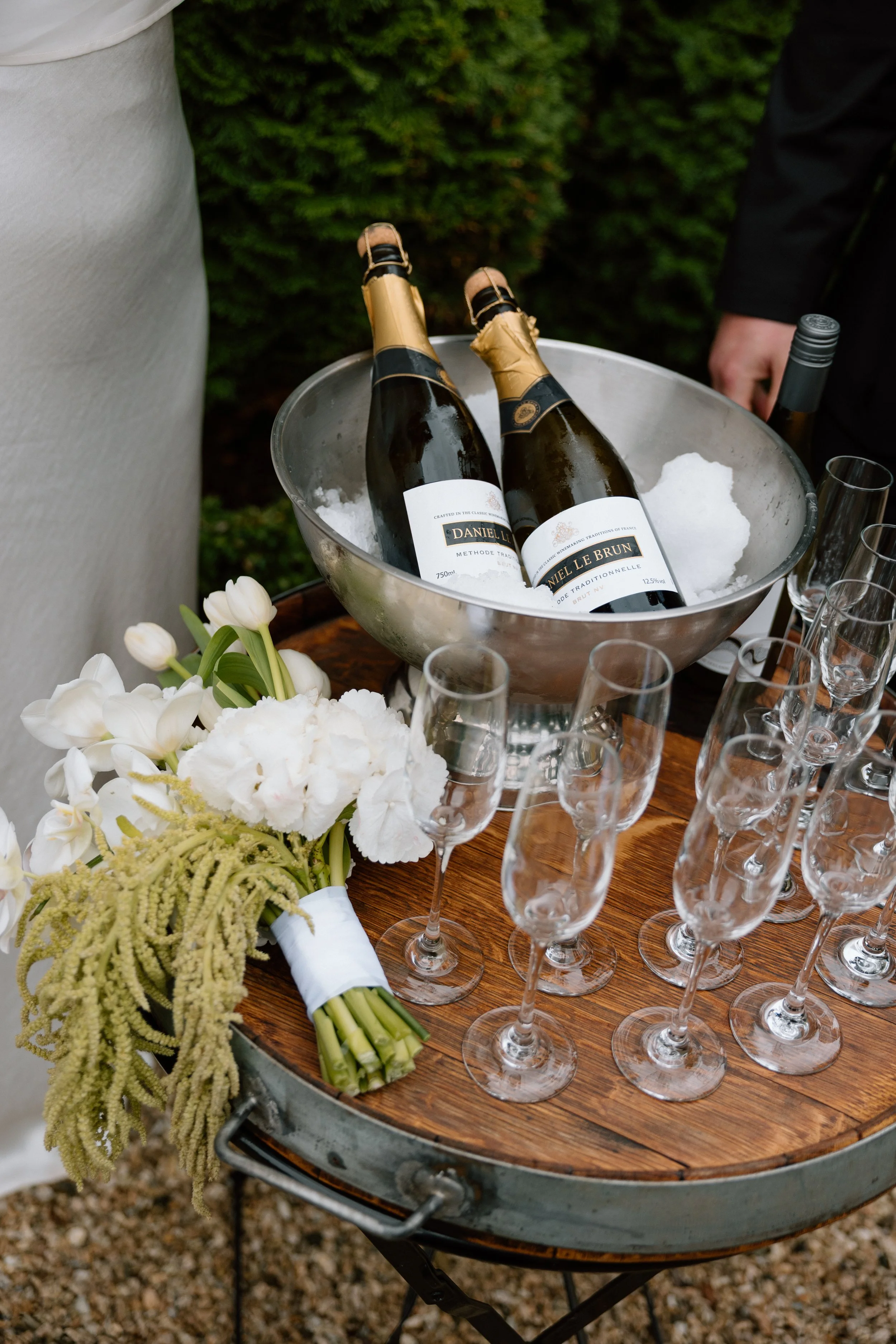 Two bottles of Daniel Le Brun sparkling wine in a silver ice bucket filled with ice, placed on a round wooden serving tray. The tray also has a bouquet of white flowers and multiple empty champagne glasses. The background includes greenery and two pe