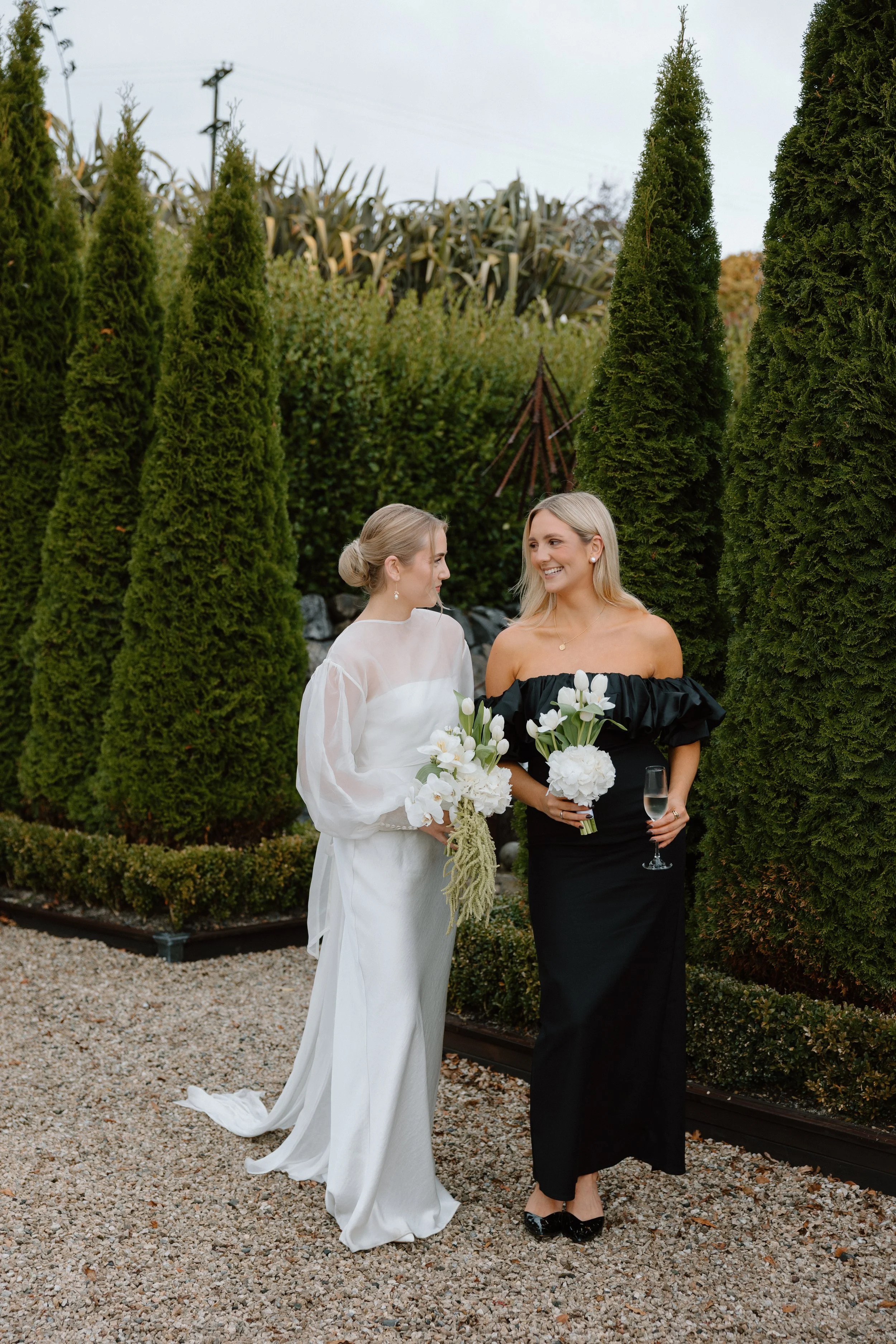 Two women standing outdoors in a garden, one in a white dress holding a bouquet, and the other in a black dress holding a glass of champagne and a bouquet, surrounded by tall green trees.