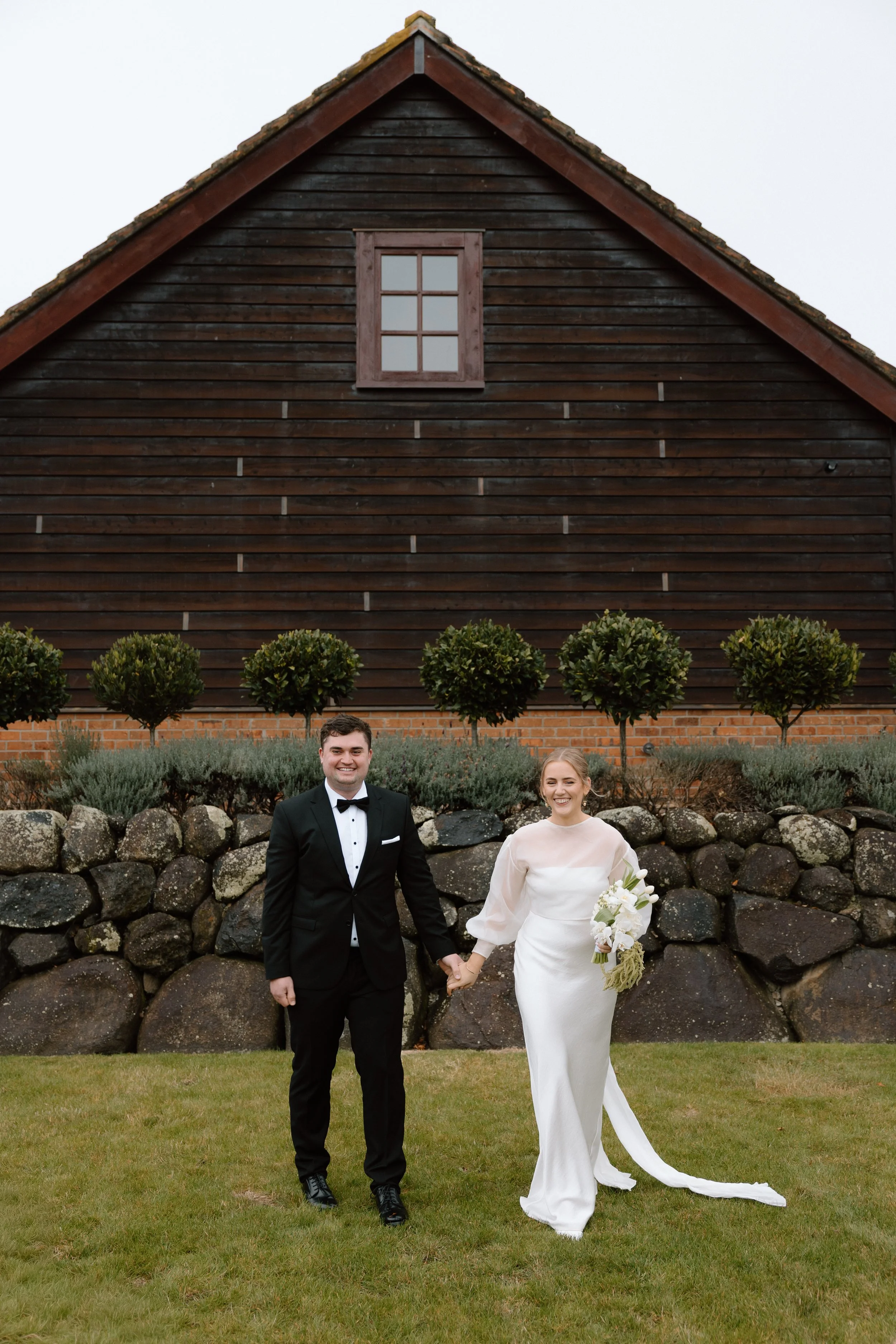 A newlywed couple holding hands outdoors in front of a stone wall with greenery and a large wooden barn in the background. The groom is dressed in a black tuxedo and the bride in a white wedding gown holding a bouquet.