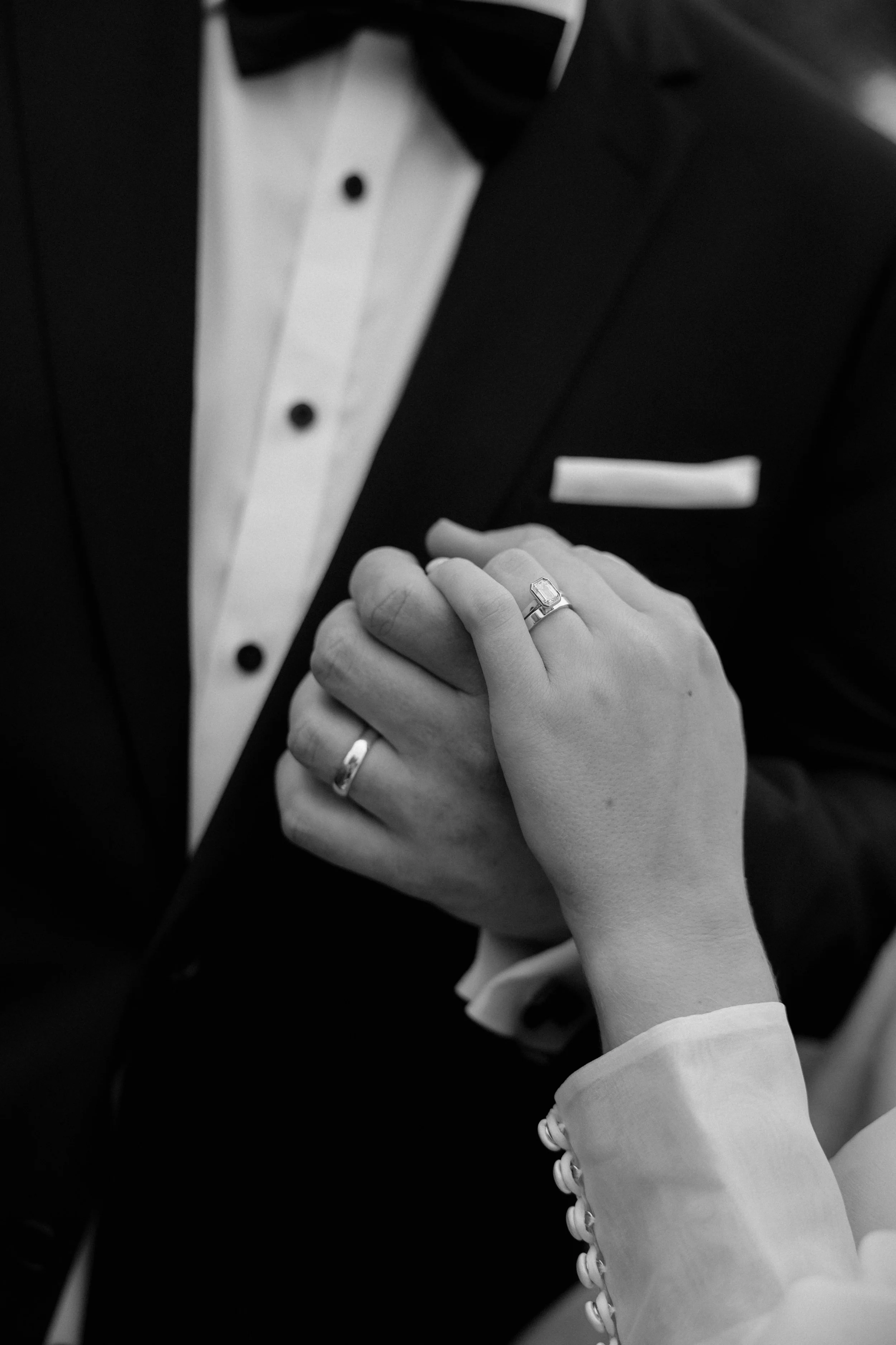 A close-up black and white photo of a person in formal attire, adjusting a tuxedo or suit with a white shirt and a black jacket. The person is wearing a ring on their ring finger and a bracelet on the wrist. The focus is on the hands and the jewelry.