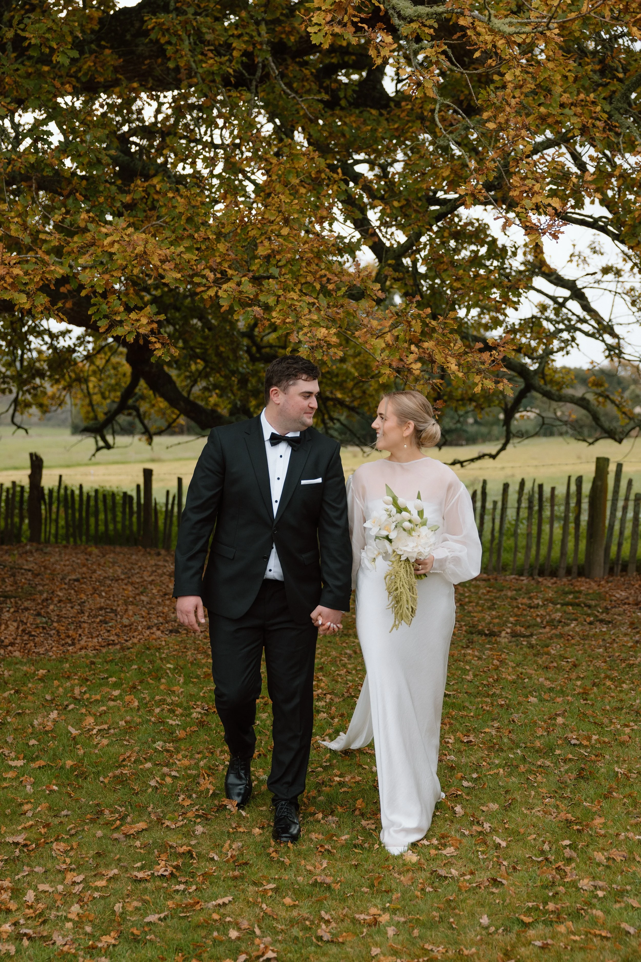A bride and groom walk hand in hand outdoors during autumn, beneath a large tree with orange and yellow leaves. The groom is dressed in a black tuxedo with a bow tie, and the bride wears a white wedding gown, holding a bouquet of white flowers. They 