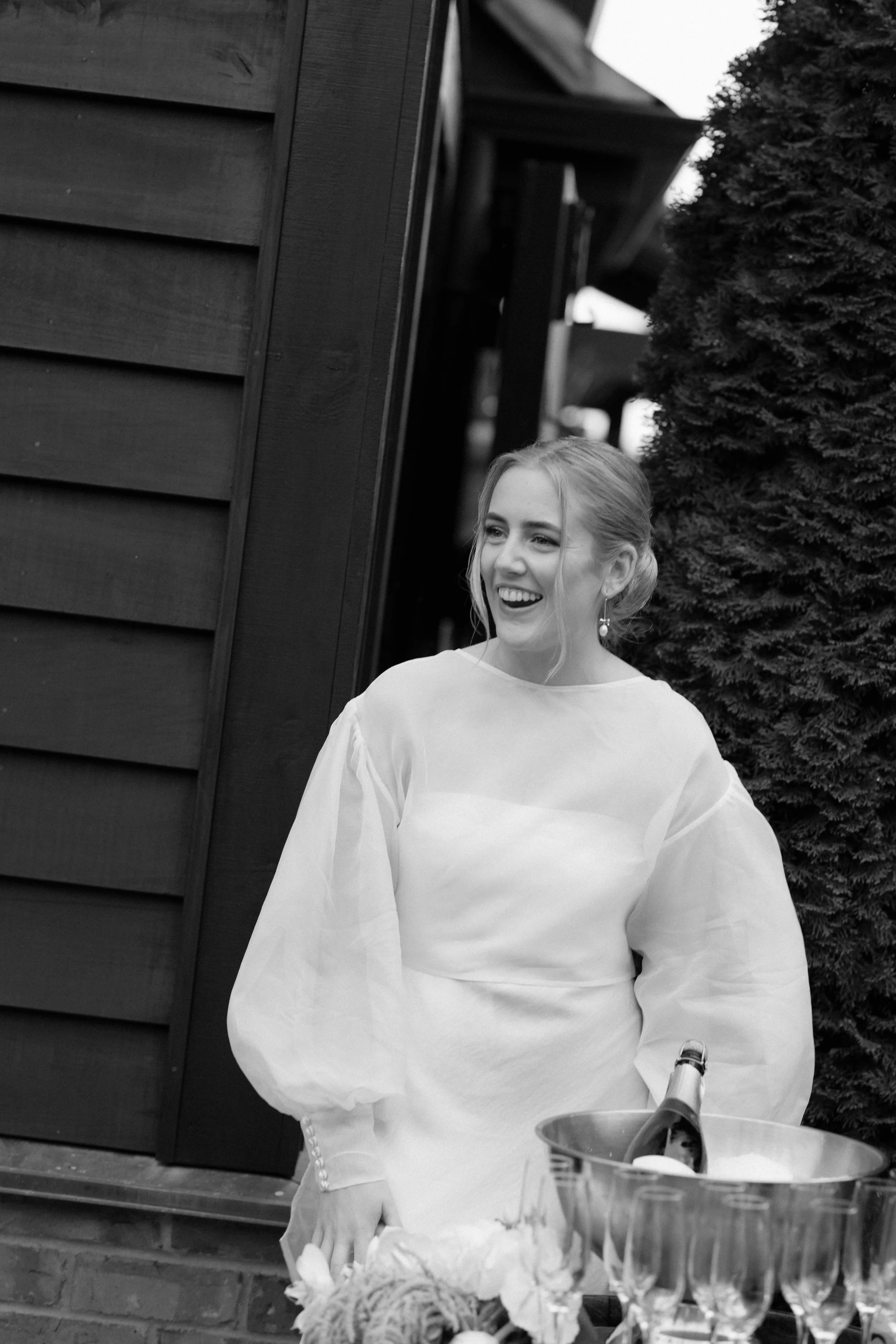 A woman smiling and holding tongs while serving food at an outdoor gathering, with wine glasses and a bottle in front of her, and a wooden house and a tree in the background.