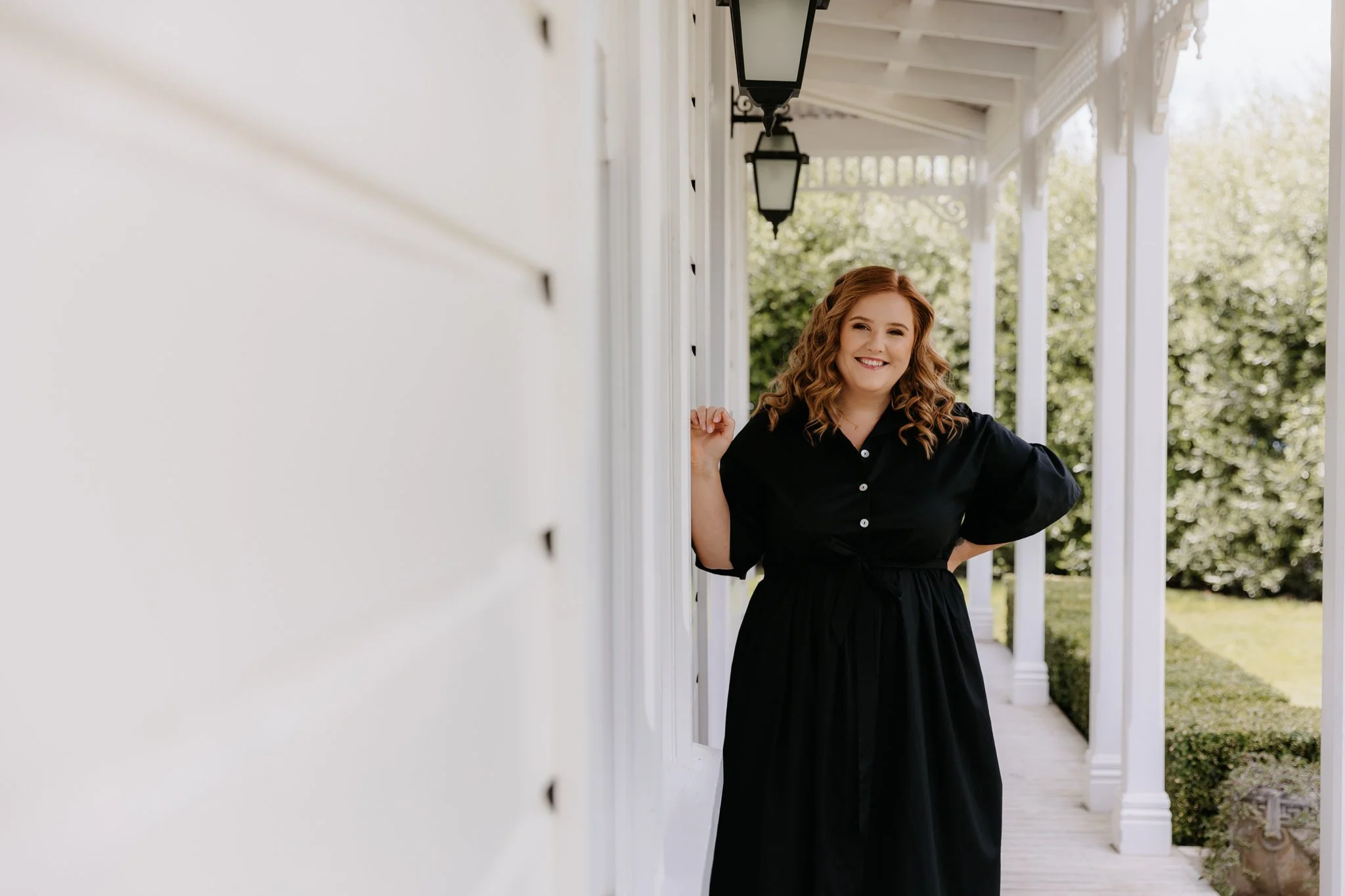 A smiling woman with curly red hair standing on a porch, leaning against a doorframe, wearing a black dress, with a background of trees and a porch with white columns and black lantern-style lights.