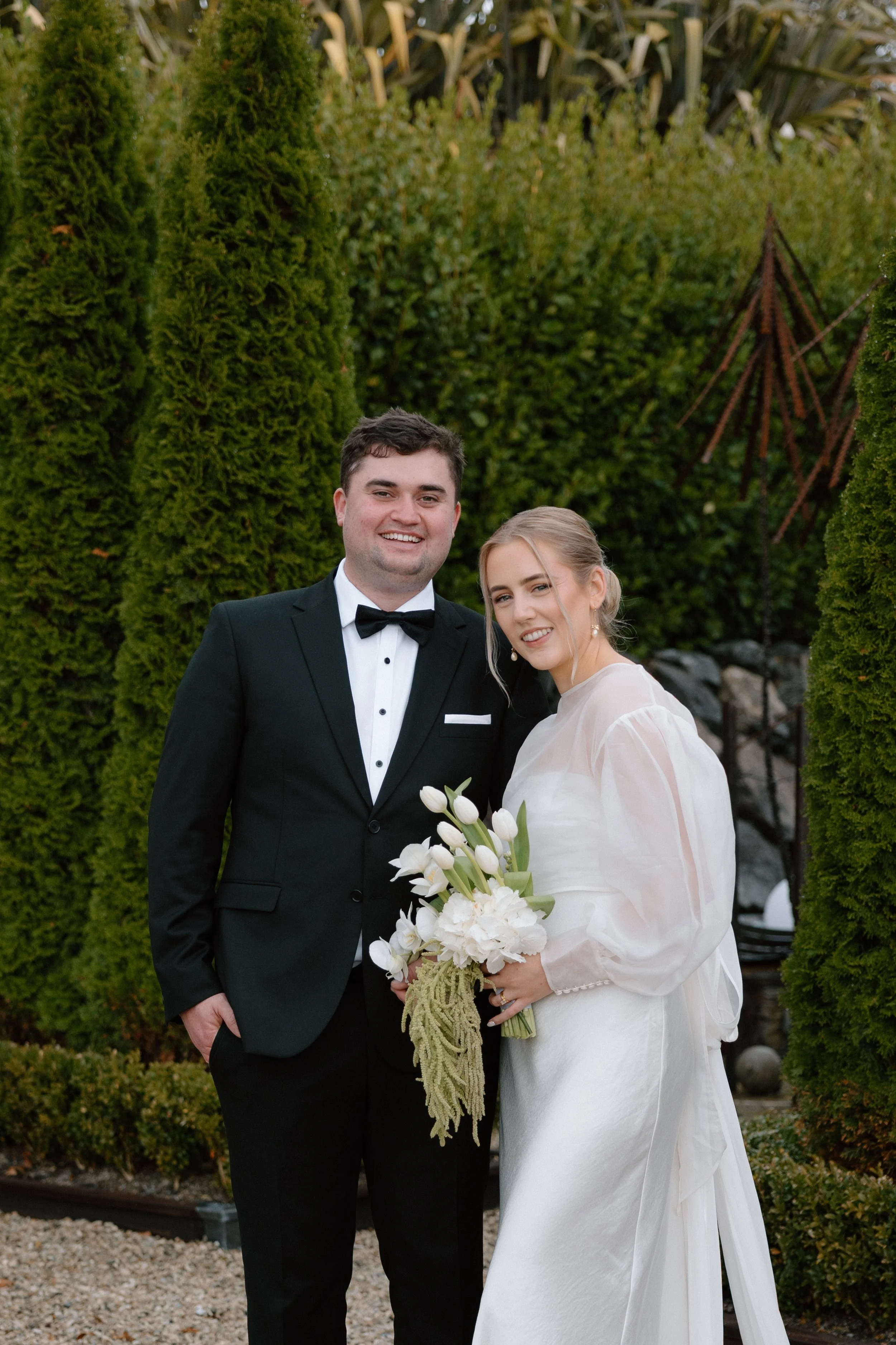 A happy newlywed couple standing outdoors among green trees and shrubbery, dressed in wedding attire, with the groom in a black tuxedo and the bride in a white wedding dress holding a bouquet of white flowers.