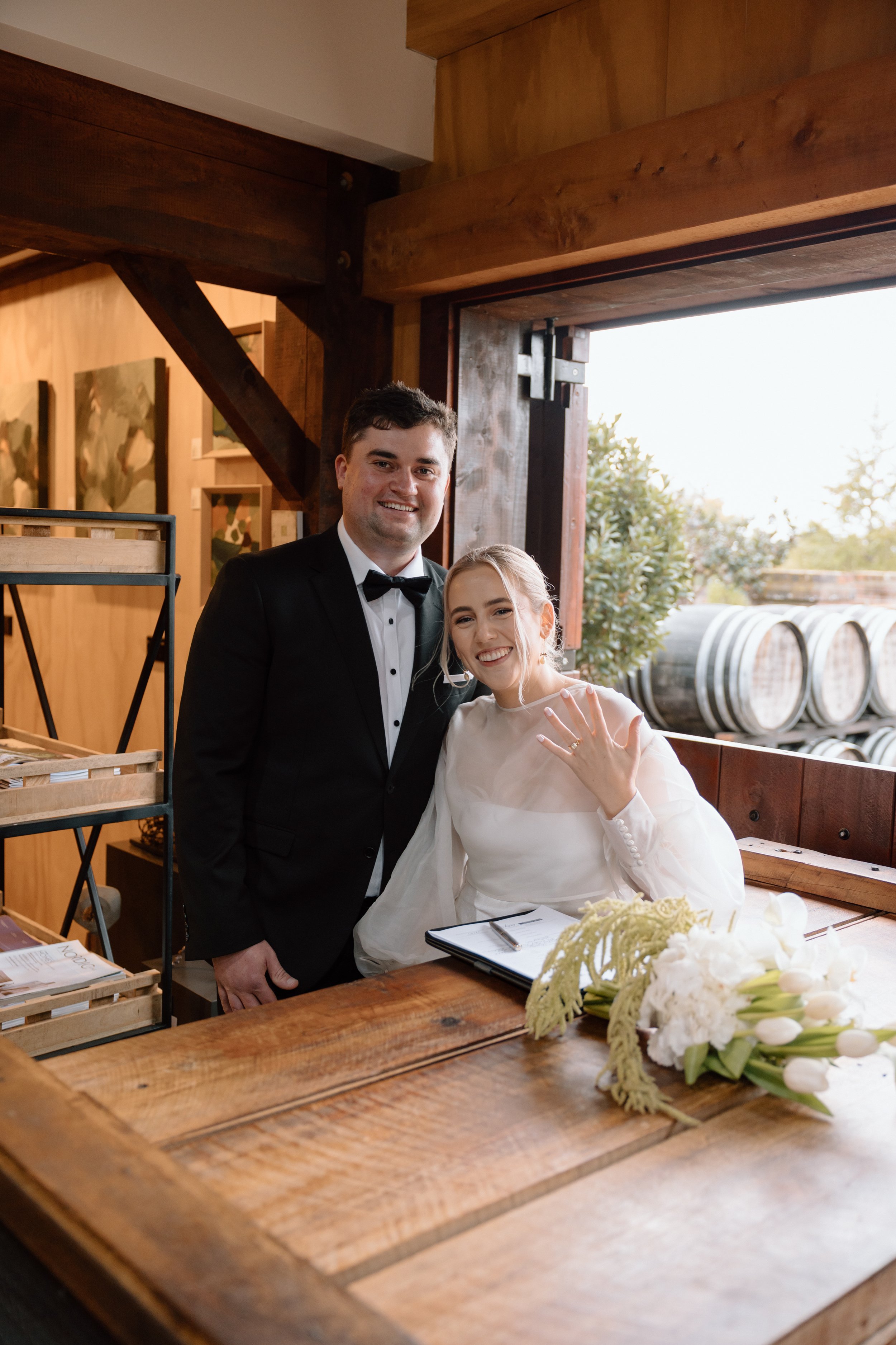 A couple dressed in wedding attire, with the groom in a tuxedo and the bride in a white wedding dress, posing together inside a rustic wooden venue with barrels visible outside.