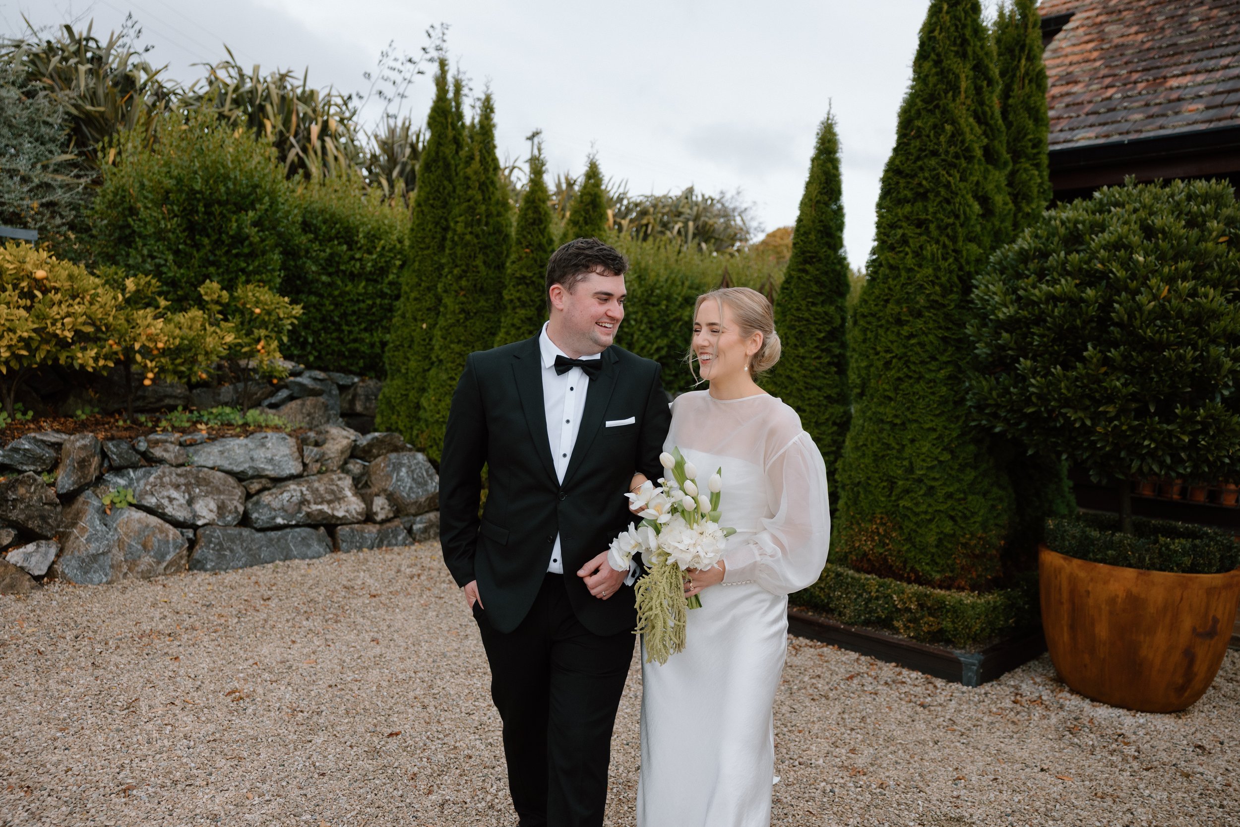 A newlywed couple smiling, walking outdoors surrounded by lush green plants and trees, with a stone wall and a large pot nearby.