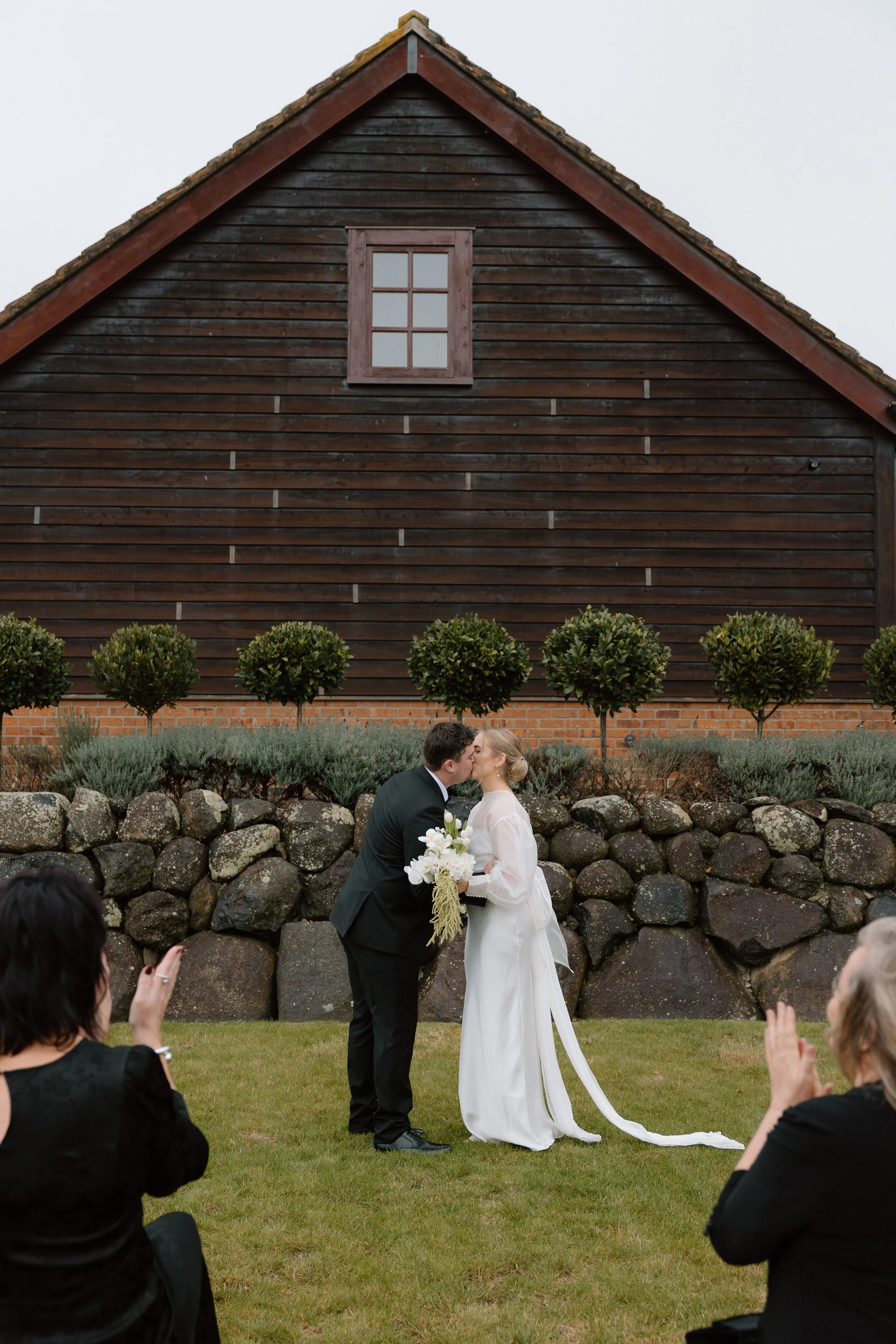 A bride and groom kissing during a wedding ceremony outdoors, with guests seated nearby and a wooden house in the background.