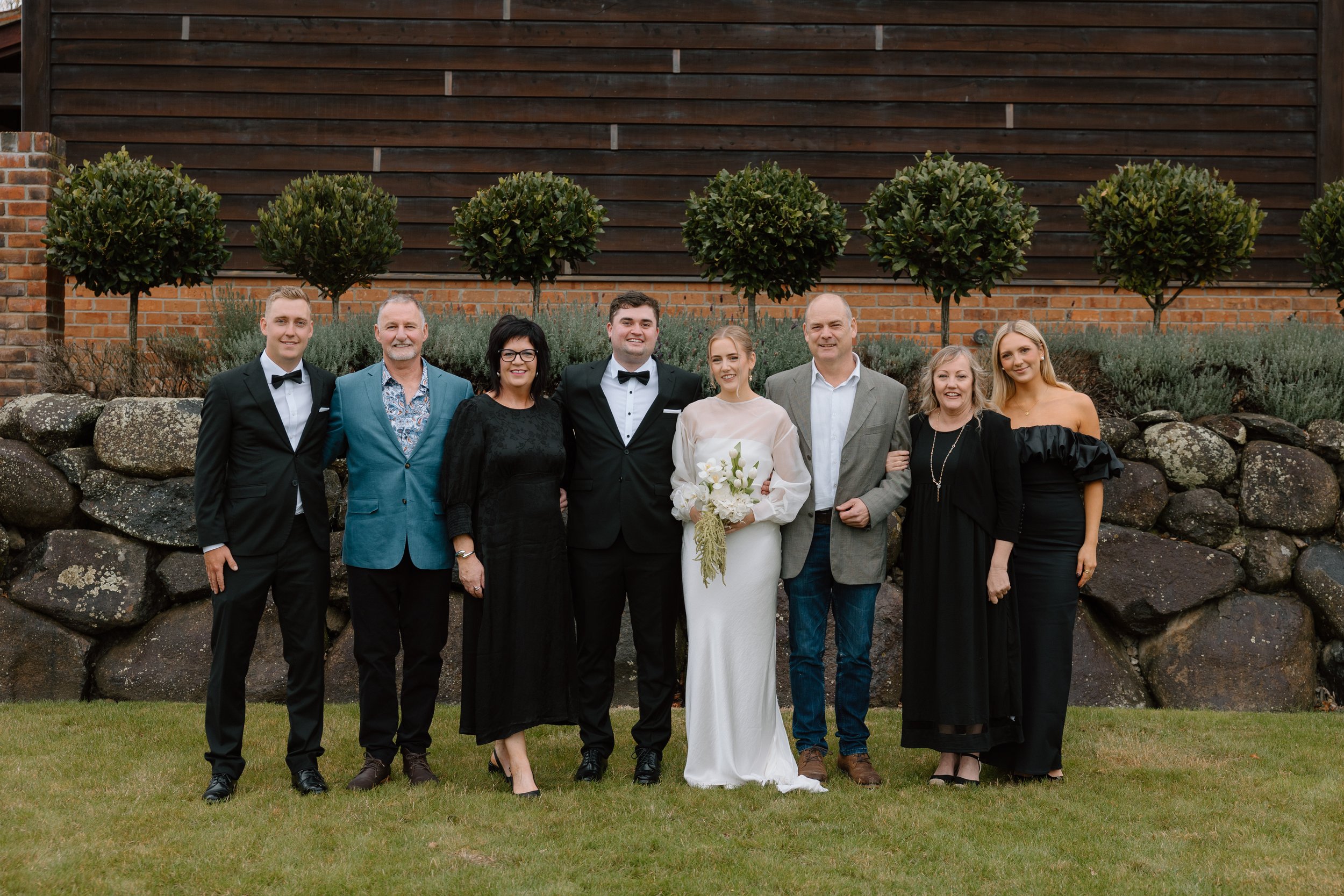 Group of eight people standing outdoors in front of a brick and wooden wall, with some greenery and rocks at the base, at a wedding or formal event.