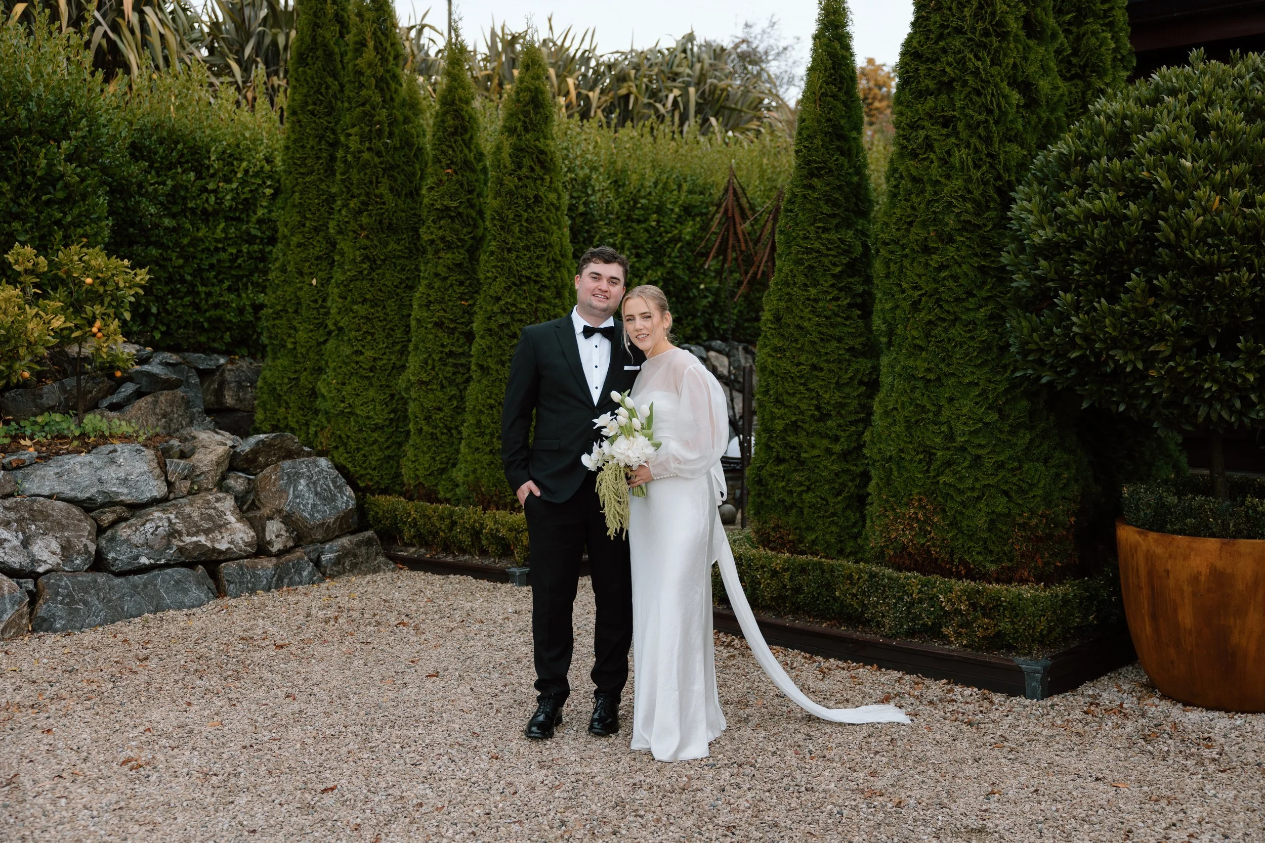 A newlywed couple standing outdoors among tall green trees and shrubs, dressed in wedding attire with the groom in a black tuxedo and the bride in a white gown holding a bouquet of white flowers.