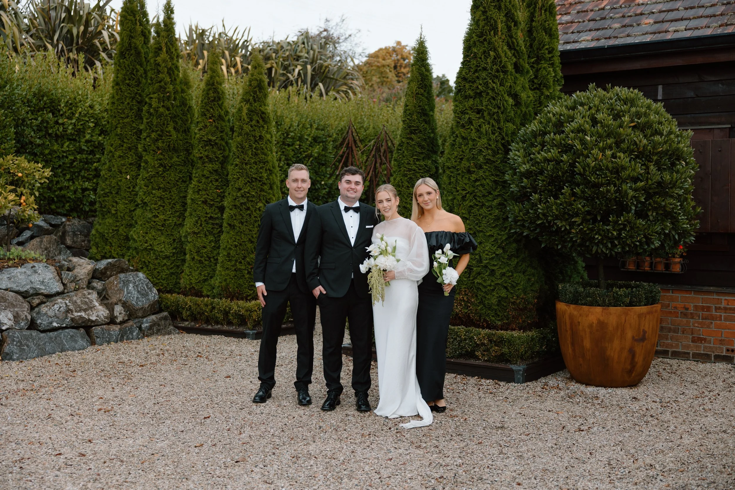 Four people dressed in formal attire, possibly at a wedding or special event, standing outdoors in front of tall evergreen trees and a dark wooden building, with a gravel and rock ground.