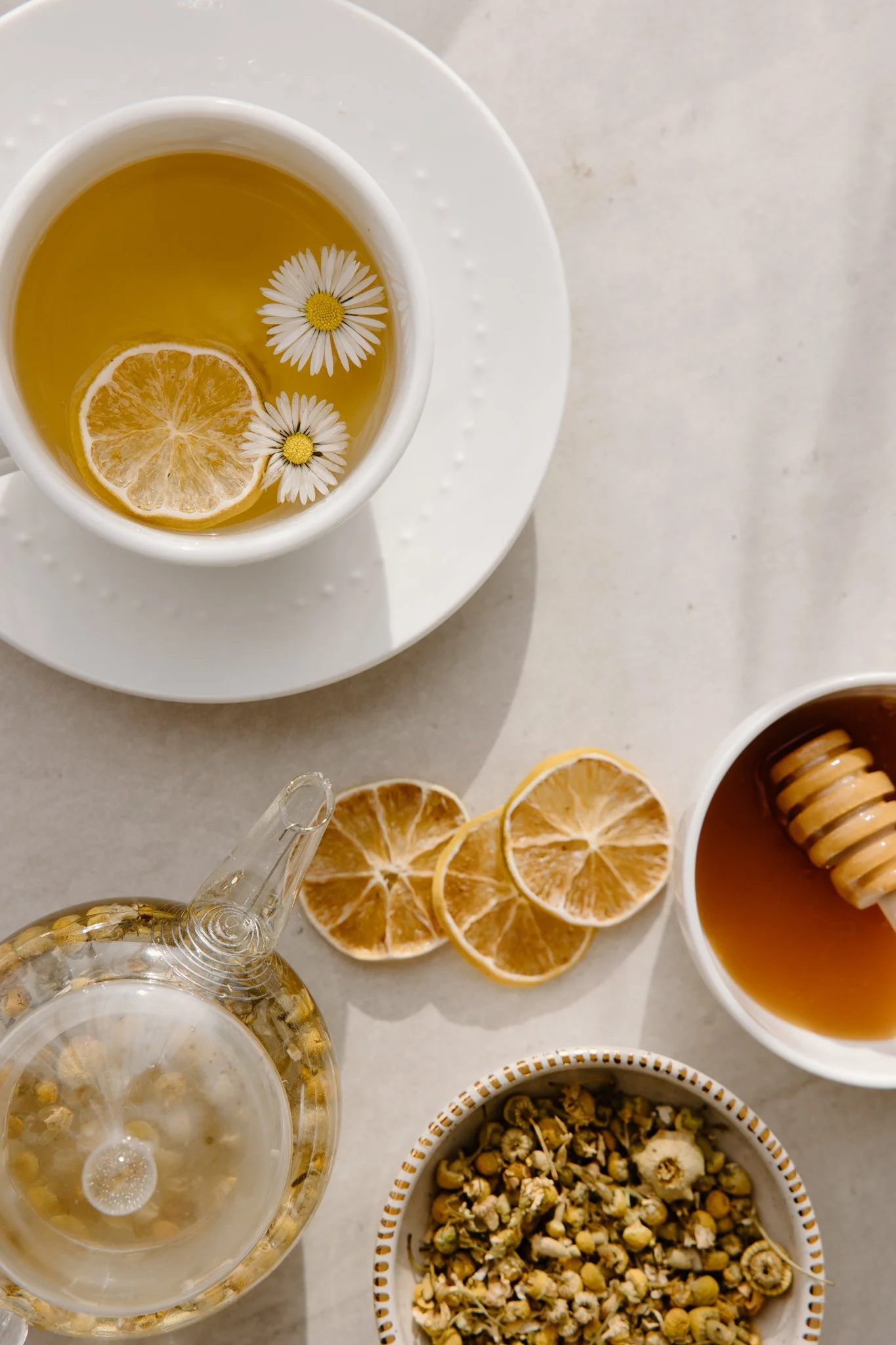 A white cup of herbal tea with lemon slice and chamomile flowers floating inside, on a white saucer, with lemon slices on the table, a glass of sparkling water, and a bowl of herbal or floral tea ingredients nearby.