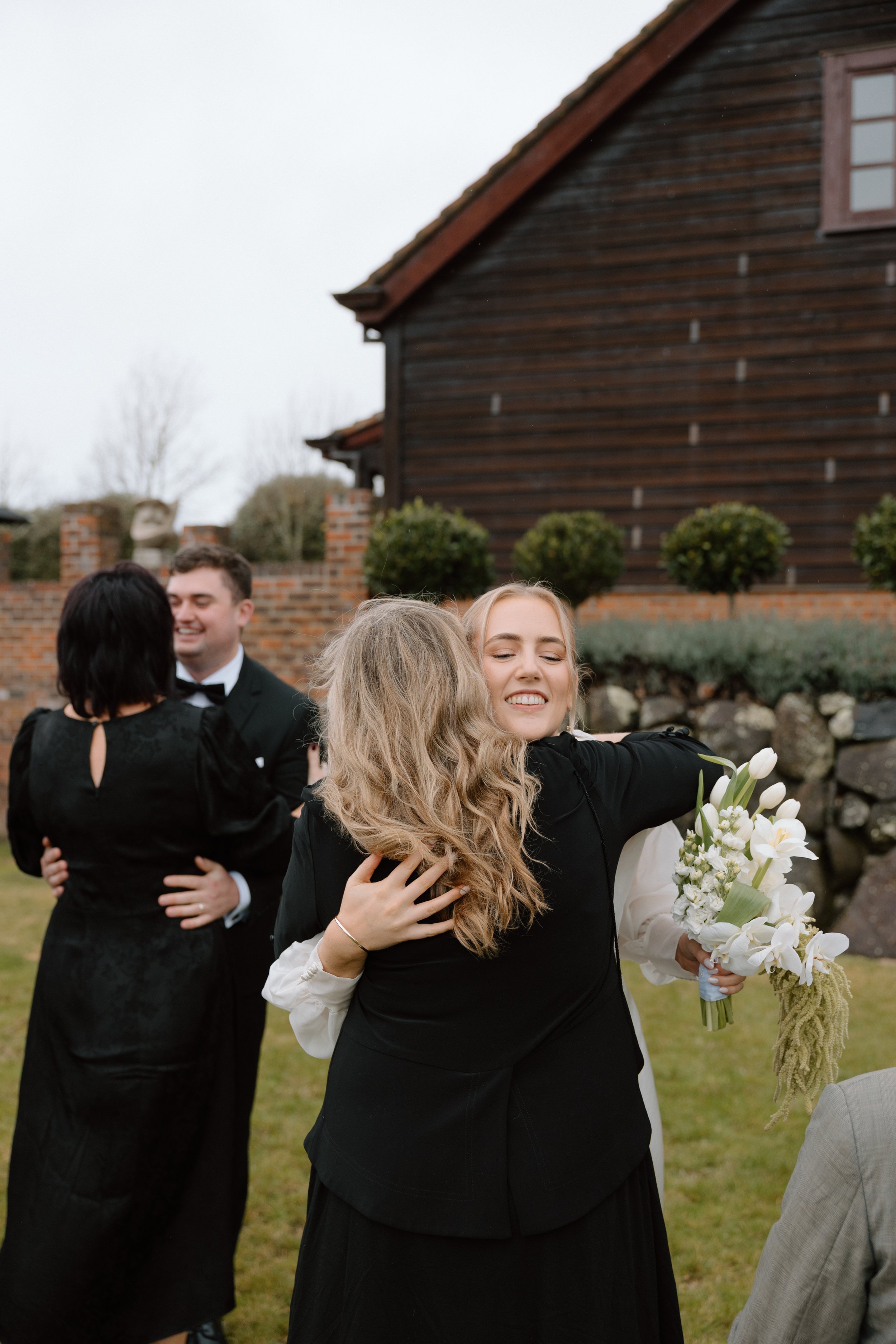 Group of people hugging outdoors in front of a house with a brick fence and trees.
