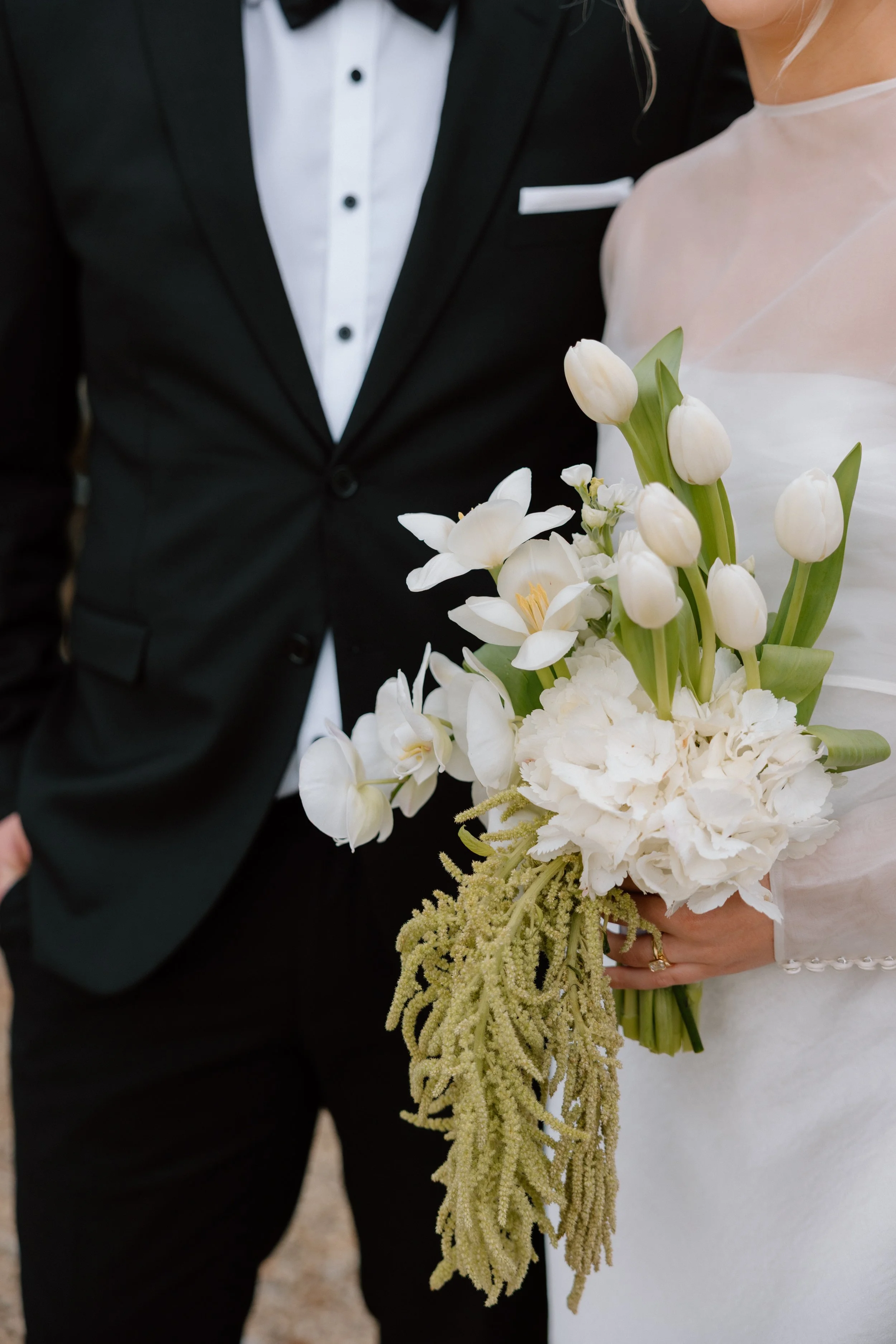 Bride standing next to groom holding a bouquet of white flowers, including tulips and orchids, at a wedding.