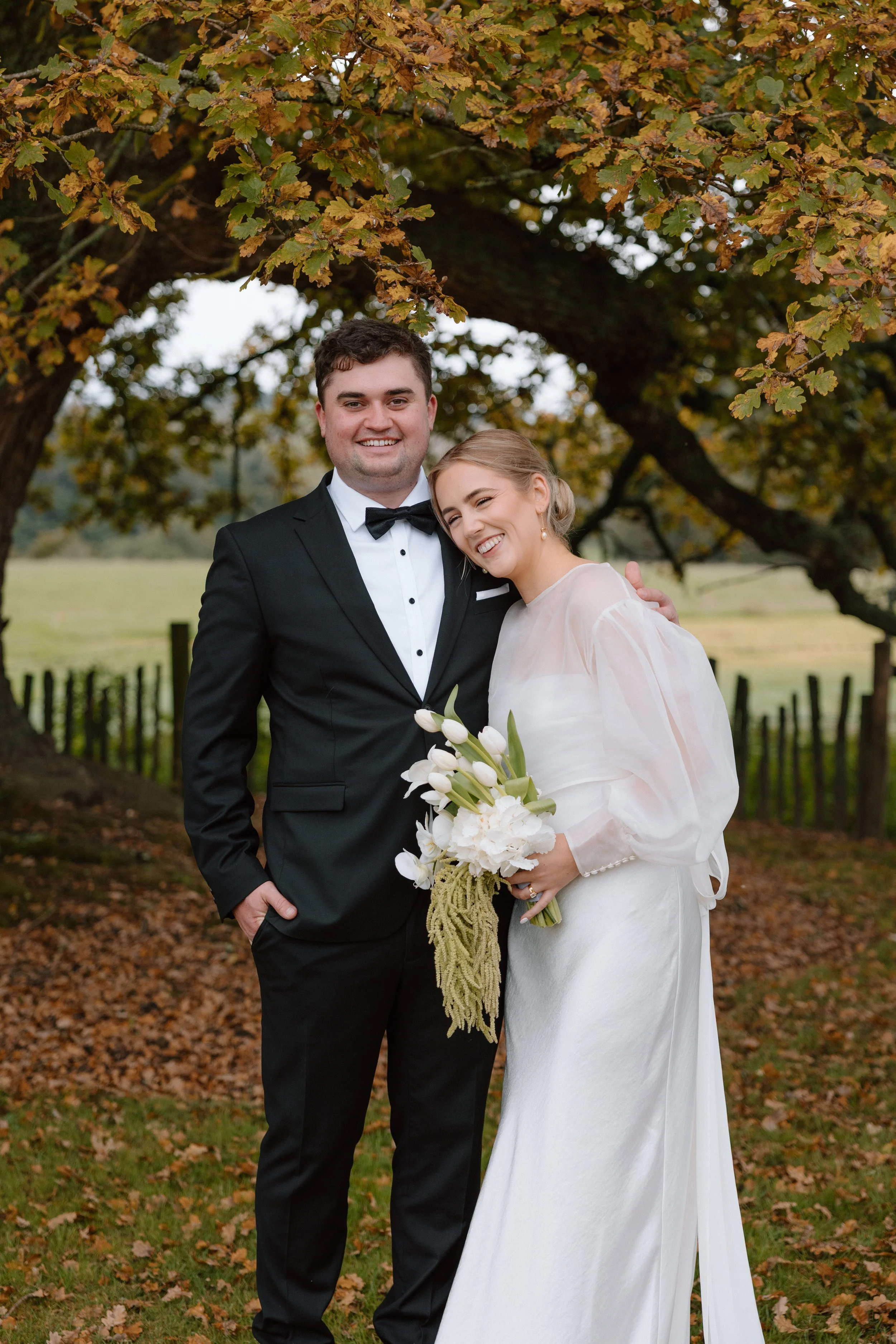 A newlywed couple standing outdoors beneath autumn leaves, smiling and embracing, with the bride holding a bouquet of white flowers.