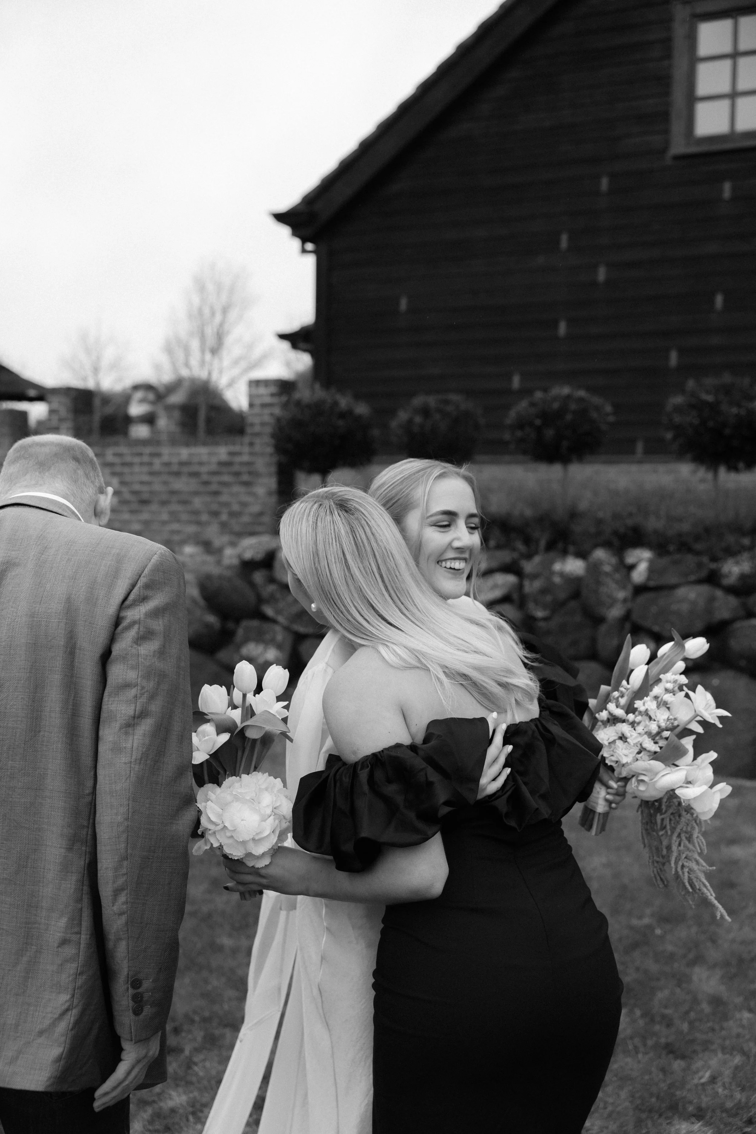 A bride and a woman embracing, holding bouquets of flowers, outdoors in front of a stone wall and a wooden house, in black and white.