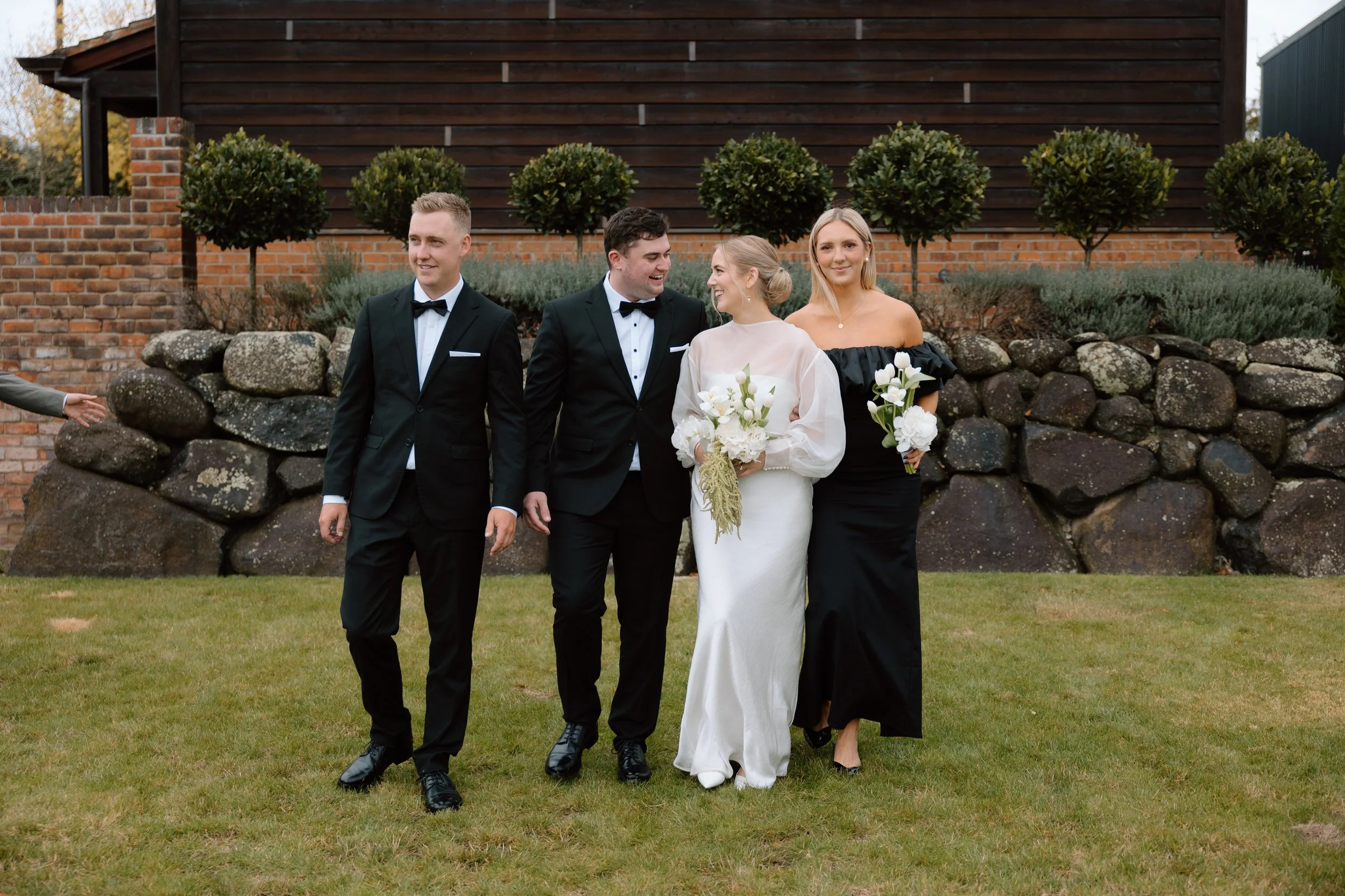 Group of four people walking outdoors in formal attire, with two men in tuxedos and two women, one in a white dress with flowers and the other in a black dress, smiling and holding bouquets, in front of a brick and wooden building with trimmed bushes
