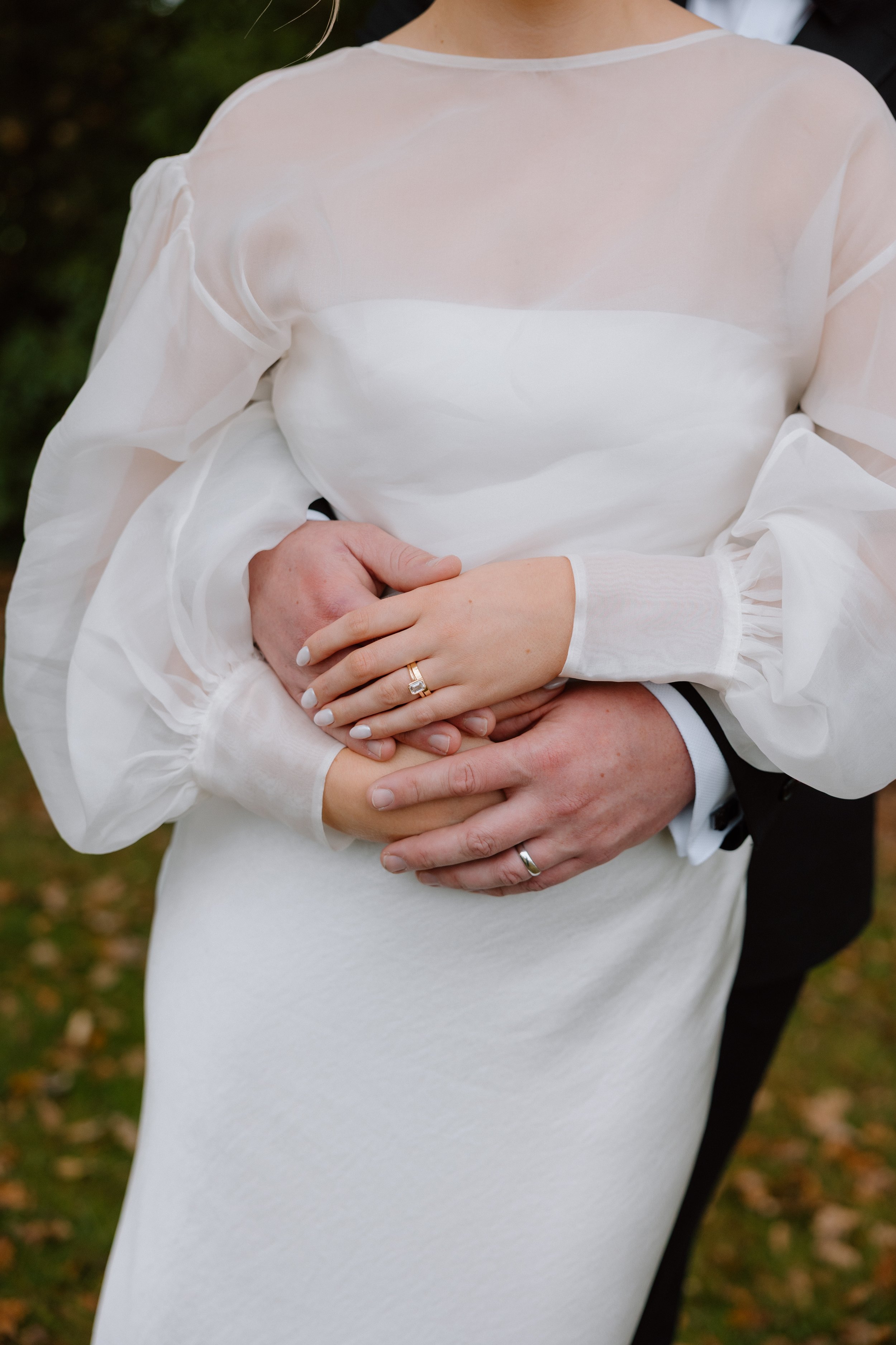 Close-up of a woman in a white dress with sheer sleeves, holding hands with a man behind her. The woman's hands rest on her abdomen with wedding rings visible. The background is outdoors with trees and fallen leaves.