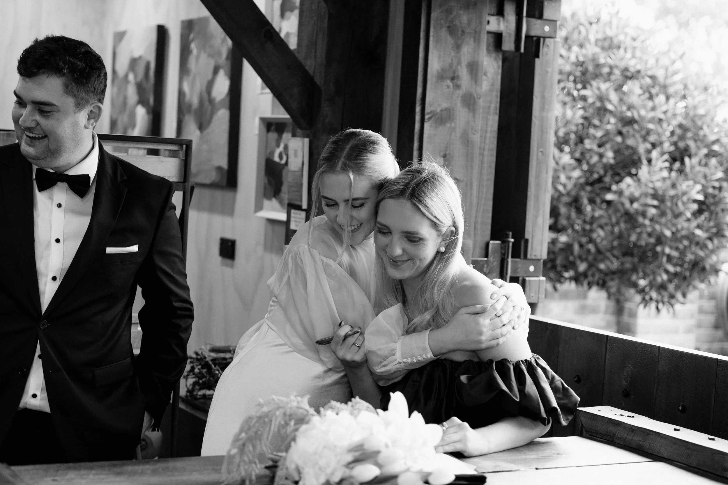 Black and white photo of a wedding reception with a bride and three guests hugging near a wooden wall, with a wedding cake on the table in the foreground.