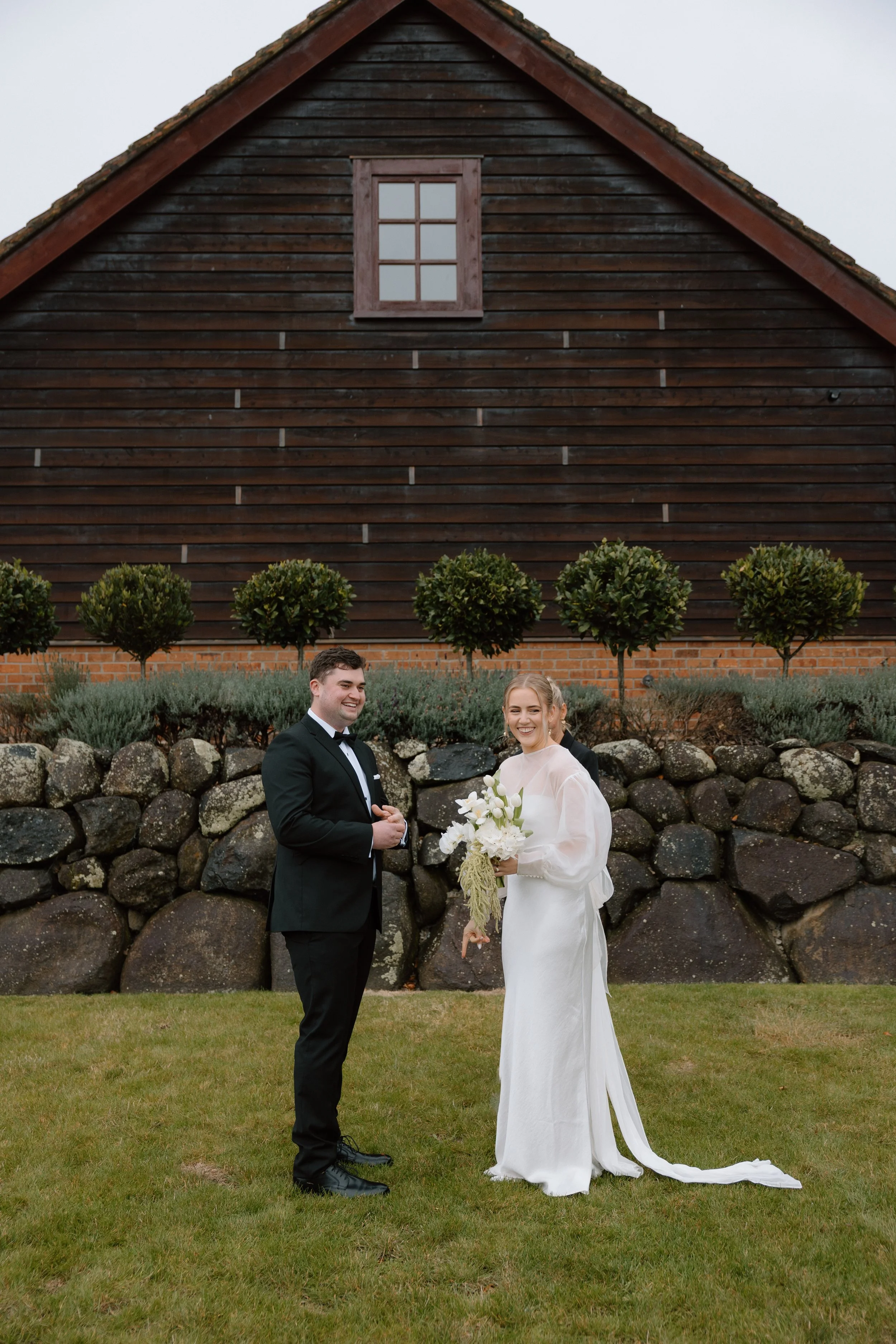 A bride and groom standing outdoors on grass, smiling at each other, with a stone wall, trees, and a large dark wooden building with a window behind them.