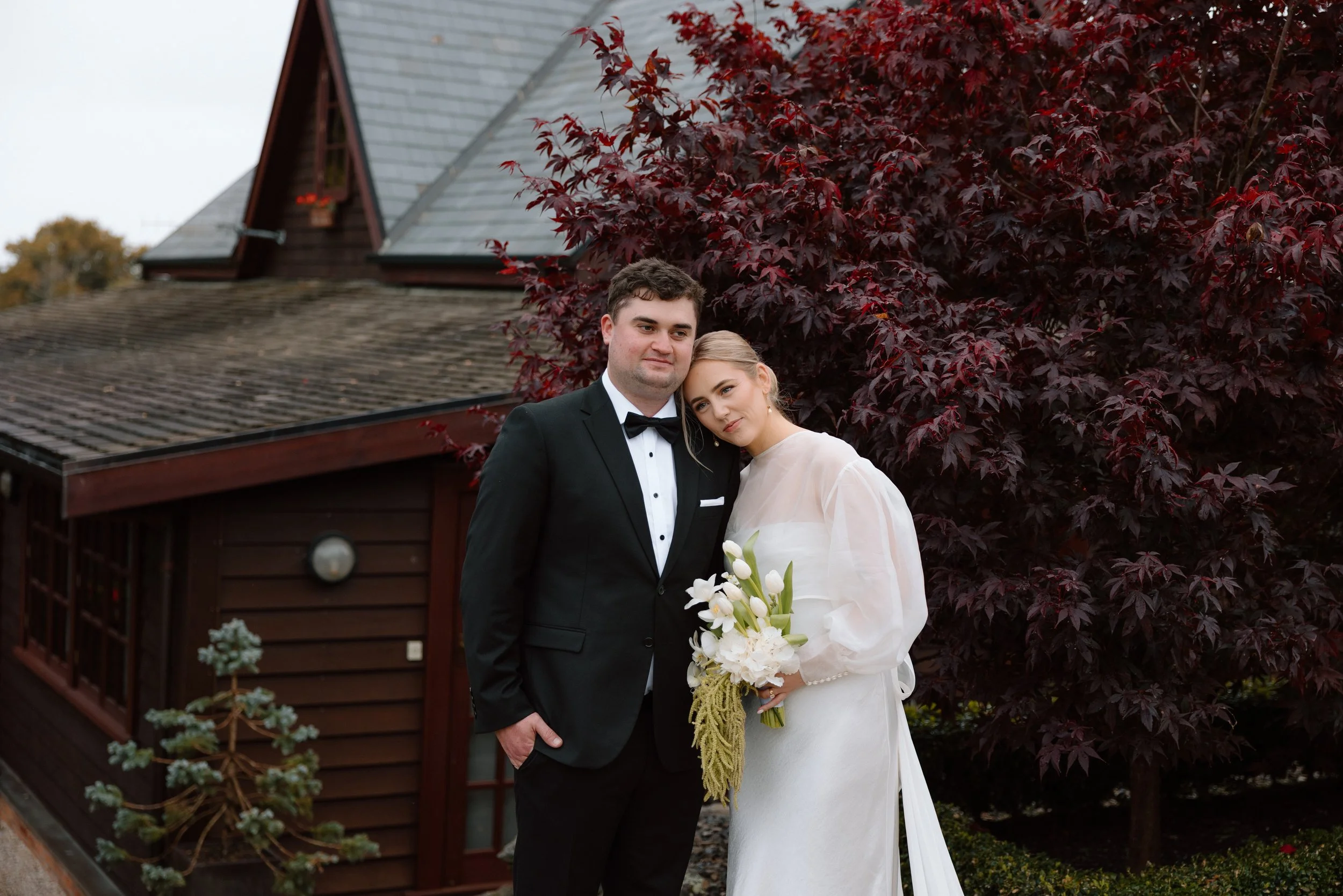 A newlywed couple standing outside in front of red-leafed trees, dressed in wedding attire. The groom is in a black tuxedo and bow tie, and the bride is in a white wedding gown holding a bouquet of white flowers.