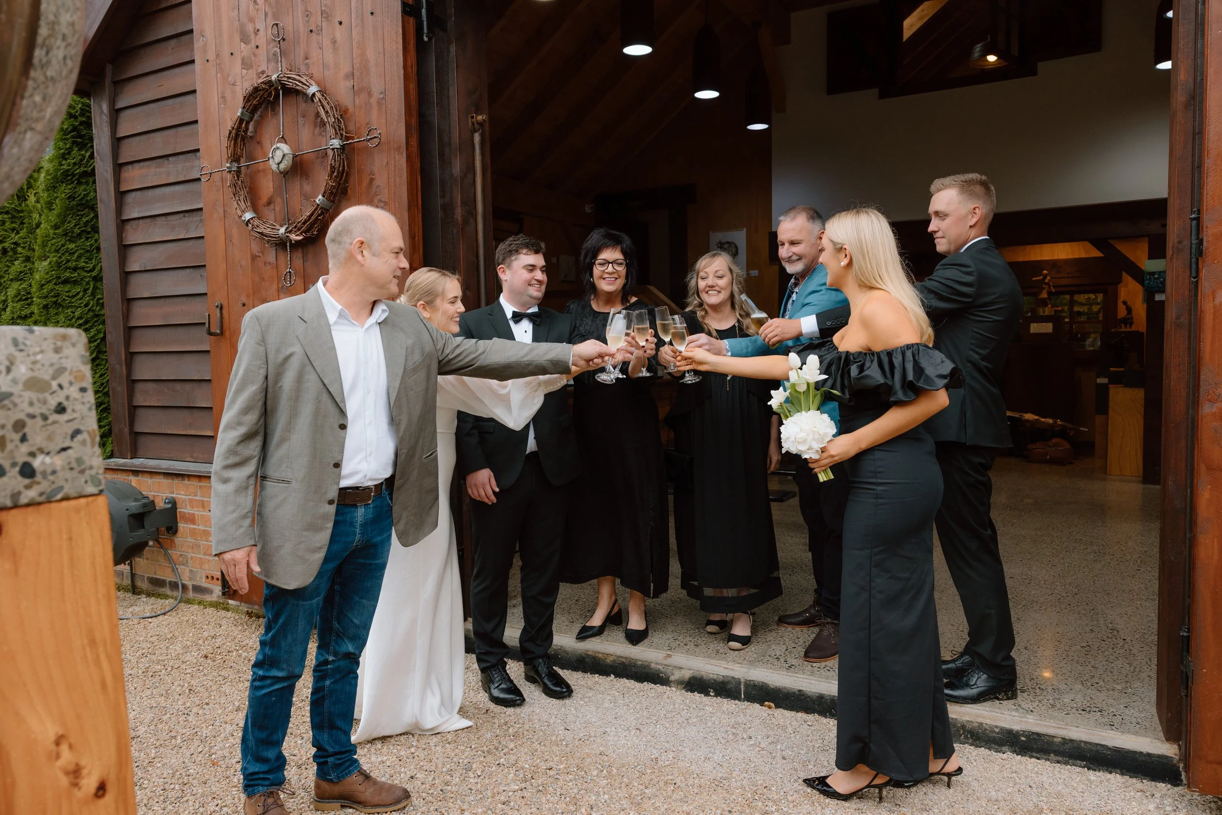 People dressed in formal wedding attire are gathered outside a rustic barn. They are toasting glasses of champagne, smiling and celebrating. A woman holds a bouquet of white flowers, and a man is wearing a light gray suit. The barn has wooden siding 