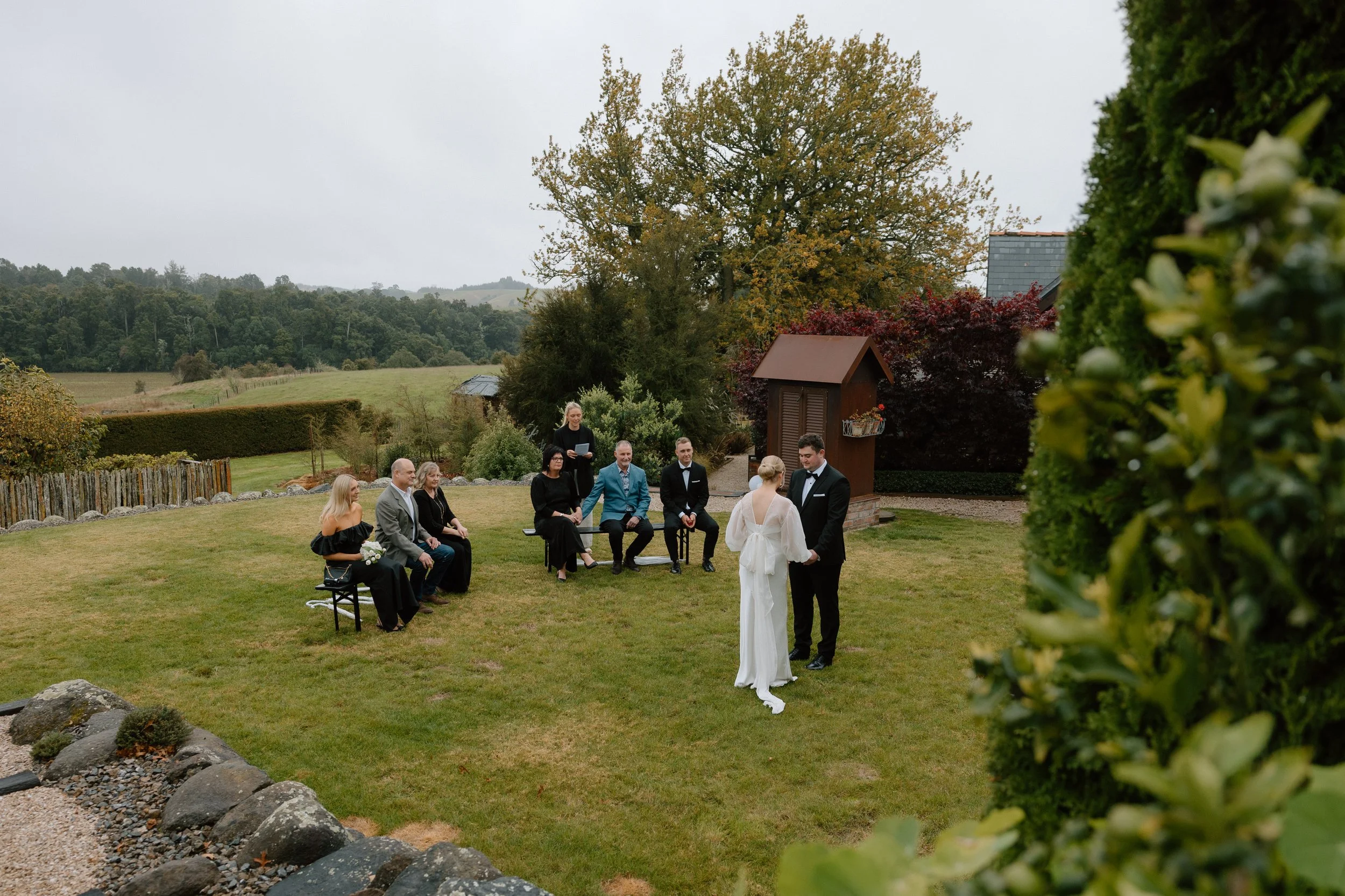 Outdoor wedding ceremony setup with brides and grooms, guests, and officiant in a scenic rural location with green fields and trees.