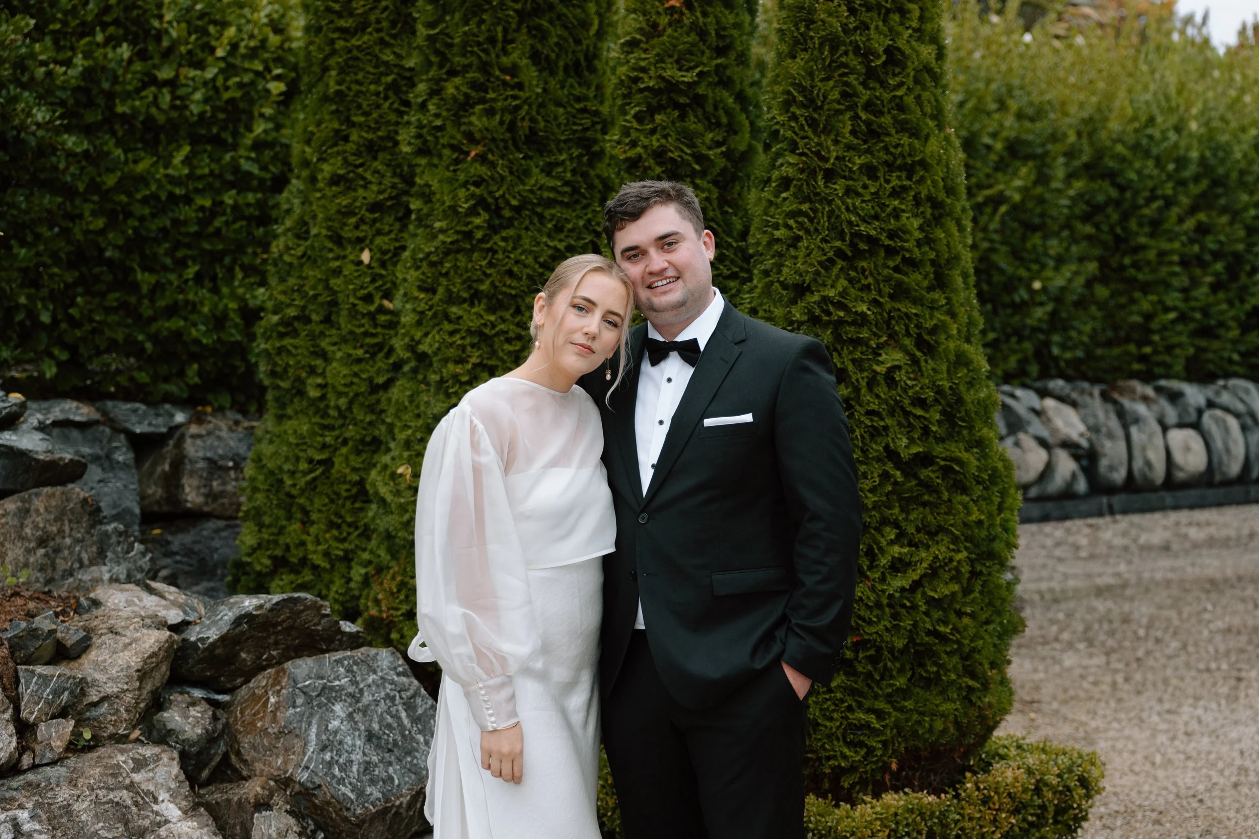 A newlywed couple standing outdoors in front of tall evergreen trees. The woman is wearing a white wedding dress and pearl earrings, and has her hair styled in an elegant updo. The man is dressed in a black tuxedo with a bowtie. They are smiling and 