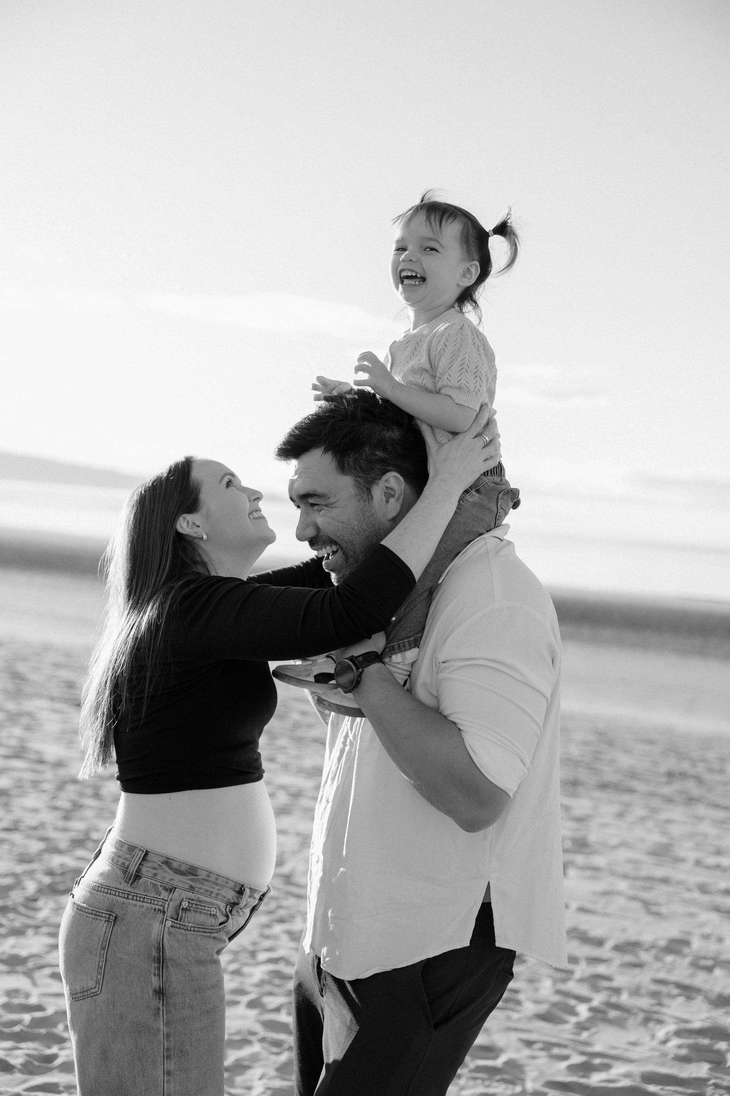 A family of three enjoying a moment together on the beach, with the mother looking happily at the father and the young girl sitting on the father's shoulders, all smiling and laughing.