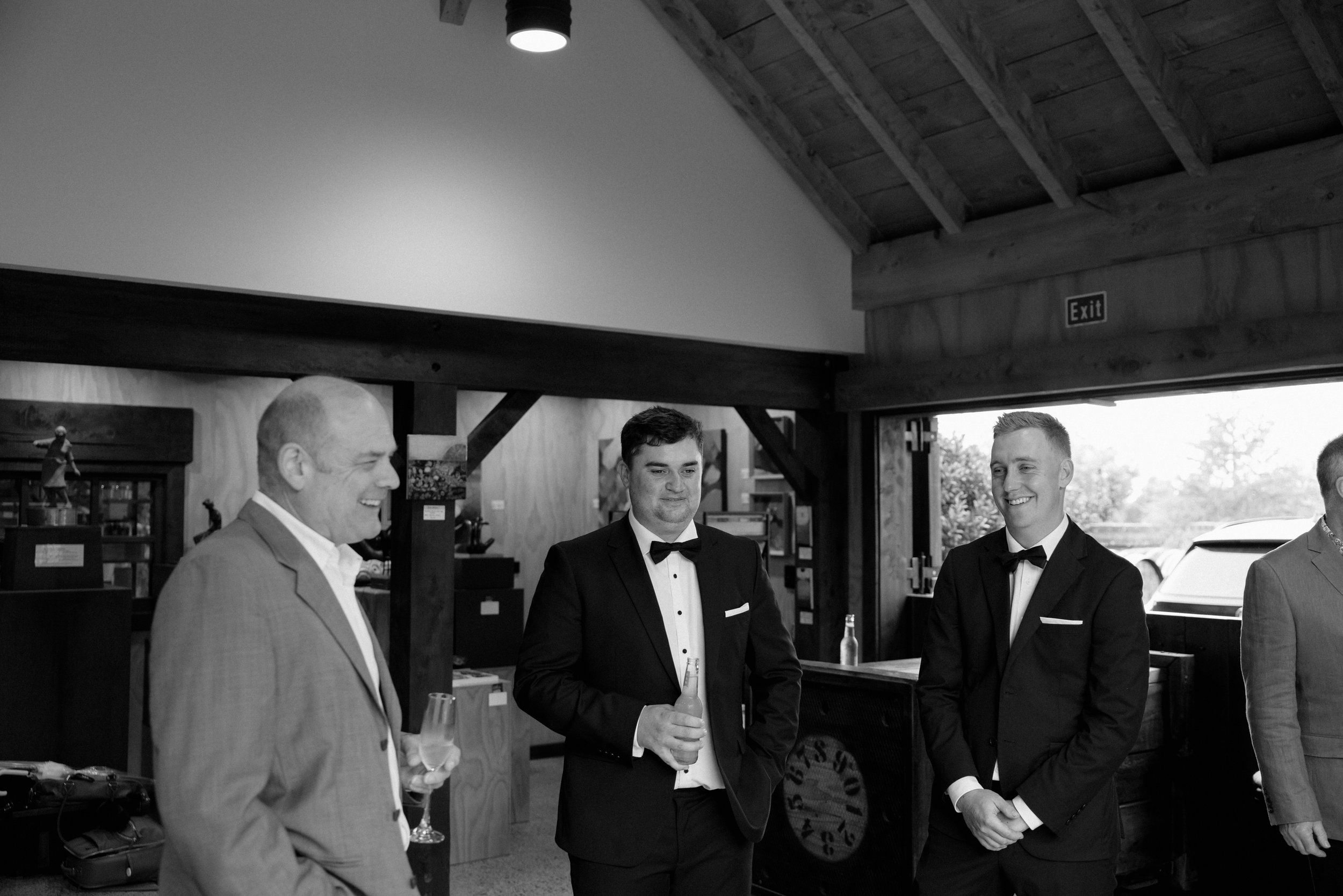 Four men dressed in tuxedos and suits are inside a rustic wooden building with high ceilings and an exit sign. They are engaged in conversation, and two of them are holding glasses of champagne.