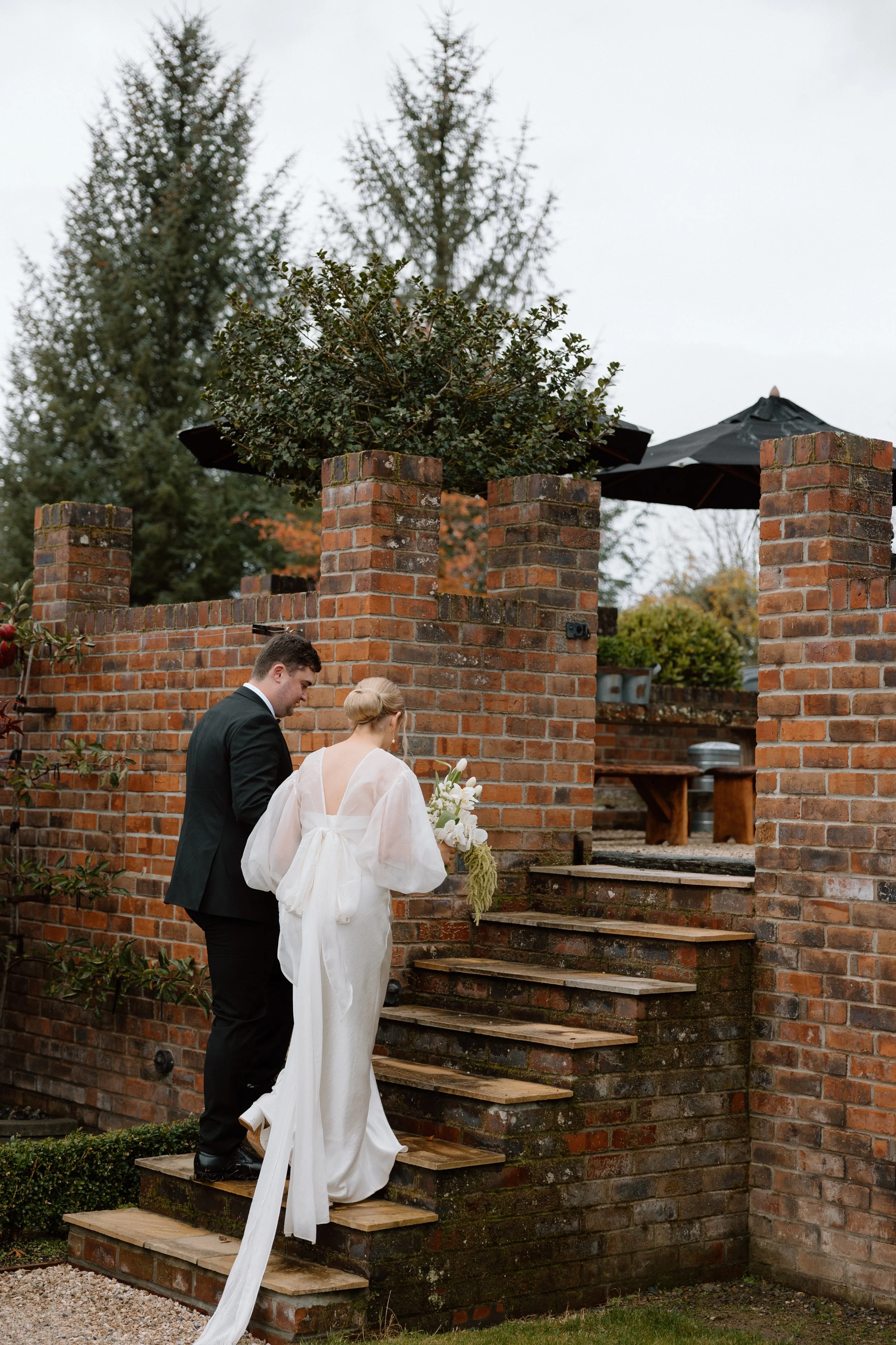 A bride and groom standing on a brick staircase outside, surrounded by a brick wall and trees, with the bride holding a bouquet of flowers.