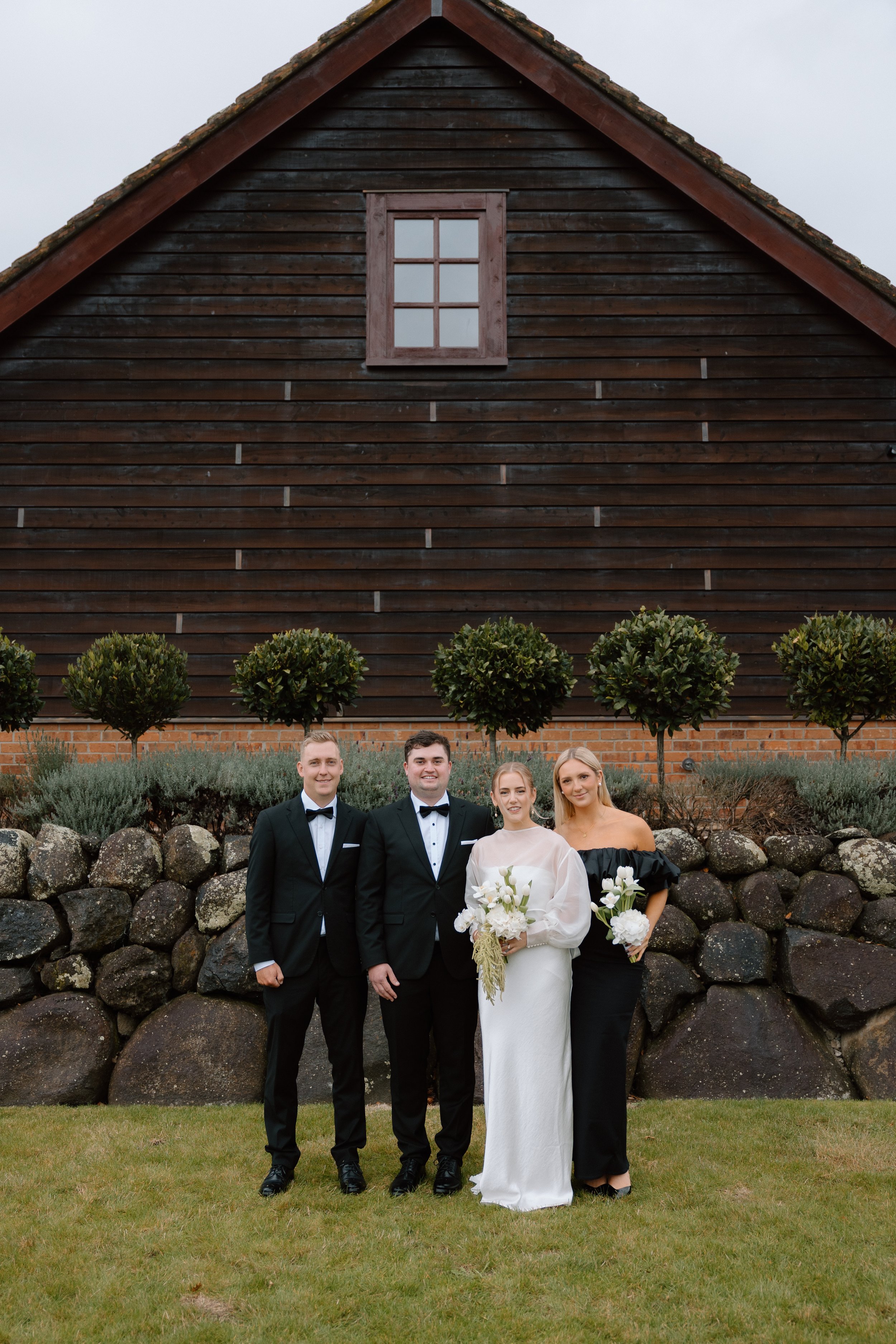 A group of four people standing outdoors in front of a stone wall, with a wooden building in the background. The group includes two men in tuxedos and two women, one in a white dress holding a bouquet and the other in a black off-shoulder dress also 
