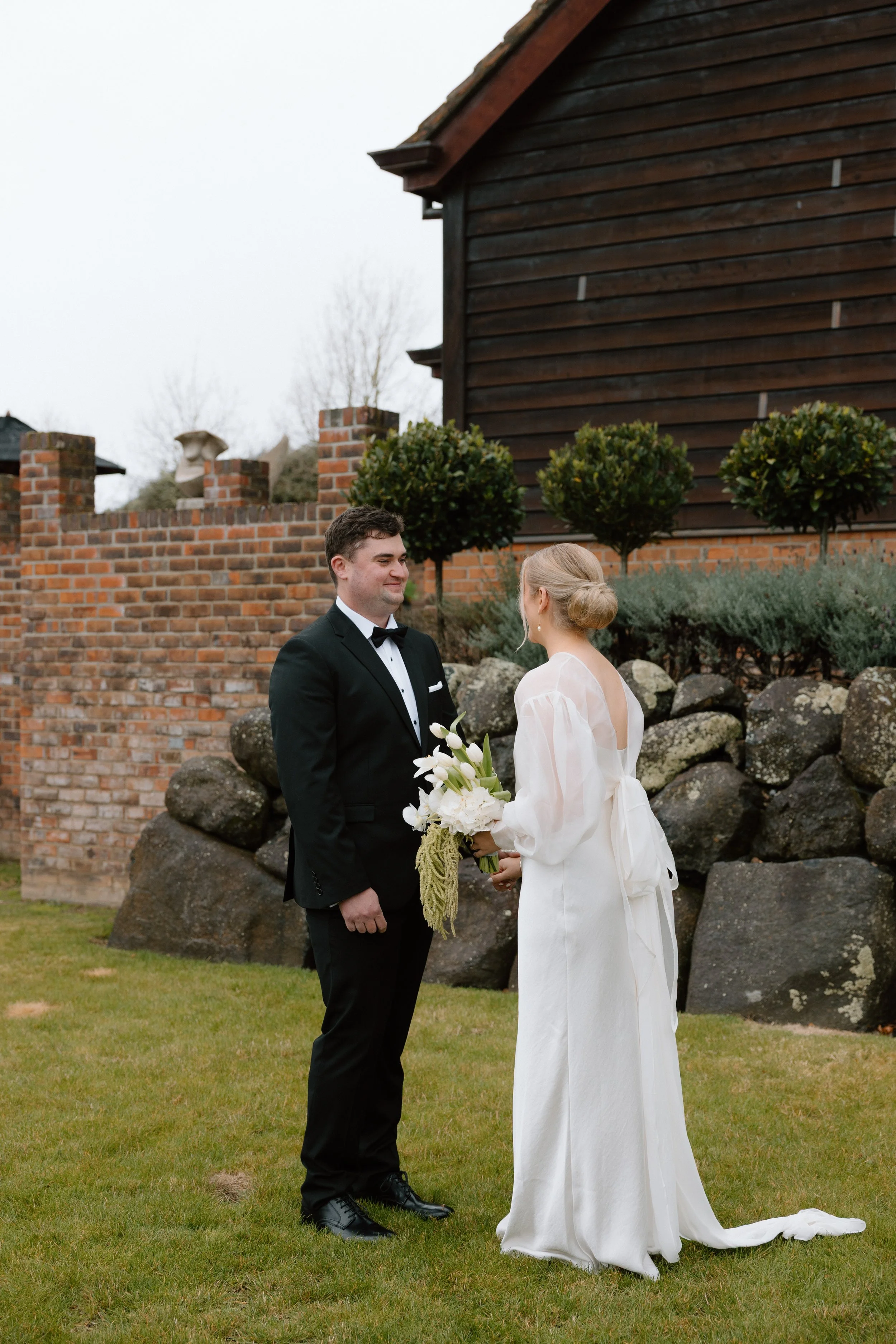 A bride and groom standing outdoors on a grassy area exchanging vows. The groom is wearing a black tuxedo and the bride is wearing a white wedding dress holding a bouquet of white flowers. Behind them is a brick wall, some small trees, and a wooden b
