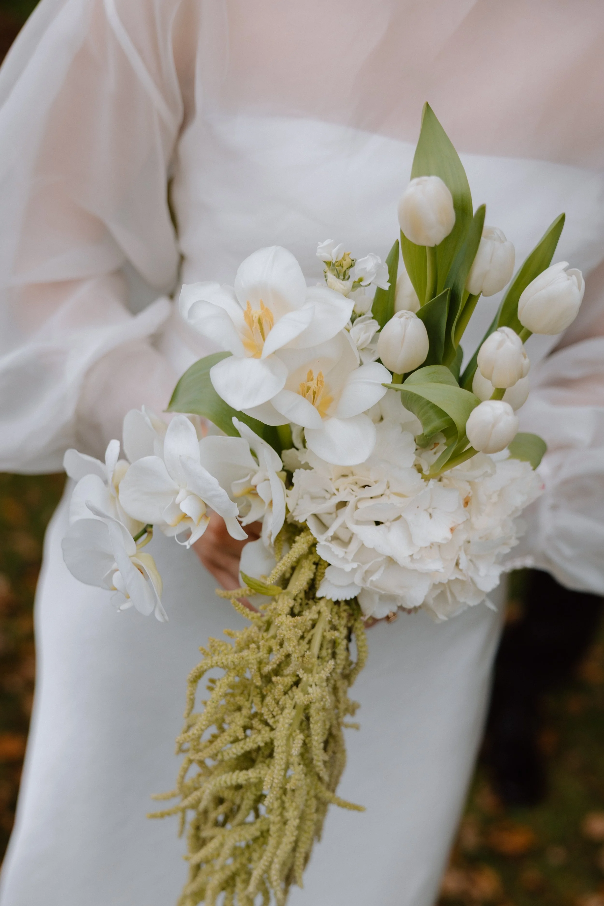 A person wearing a white garment holding a bouquet of white flowers, including tulips, orchids, and other small white flowers, with a trailing beige decorative element.