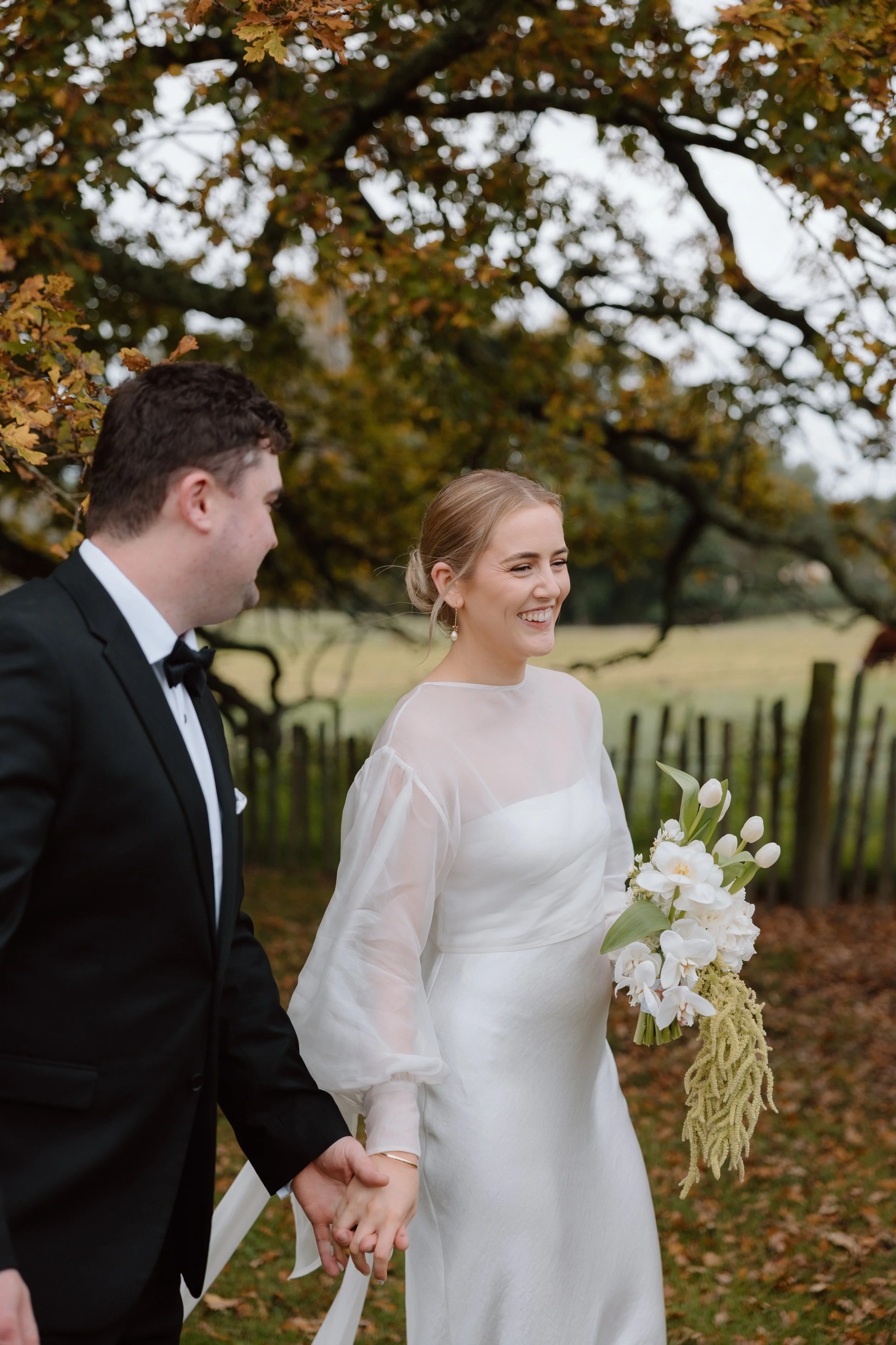 A bride and groom holding hands outdoors with trees and a wooden fence in the background. The bride is smiling and holding a bouquet of white flowers, wearing a white dress with sheer sleeves. The groom is wearing a black tuxedo with a bow tie.