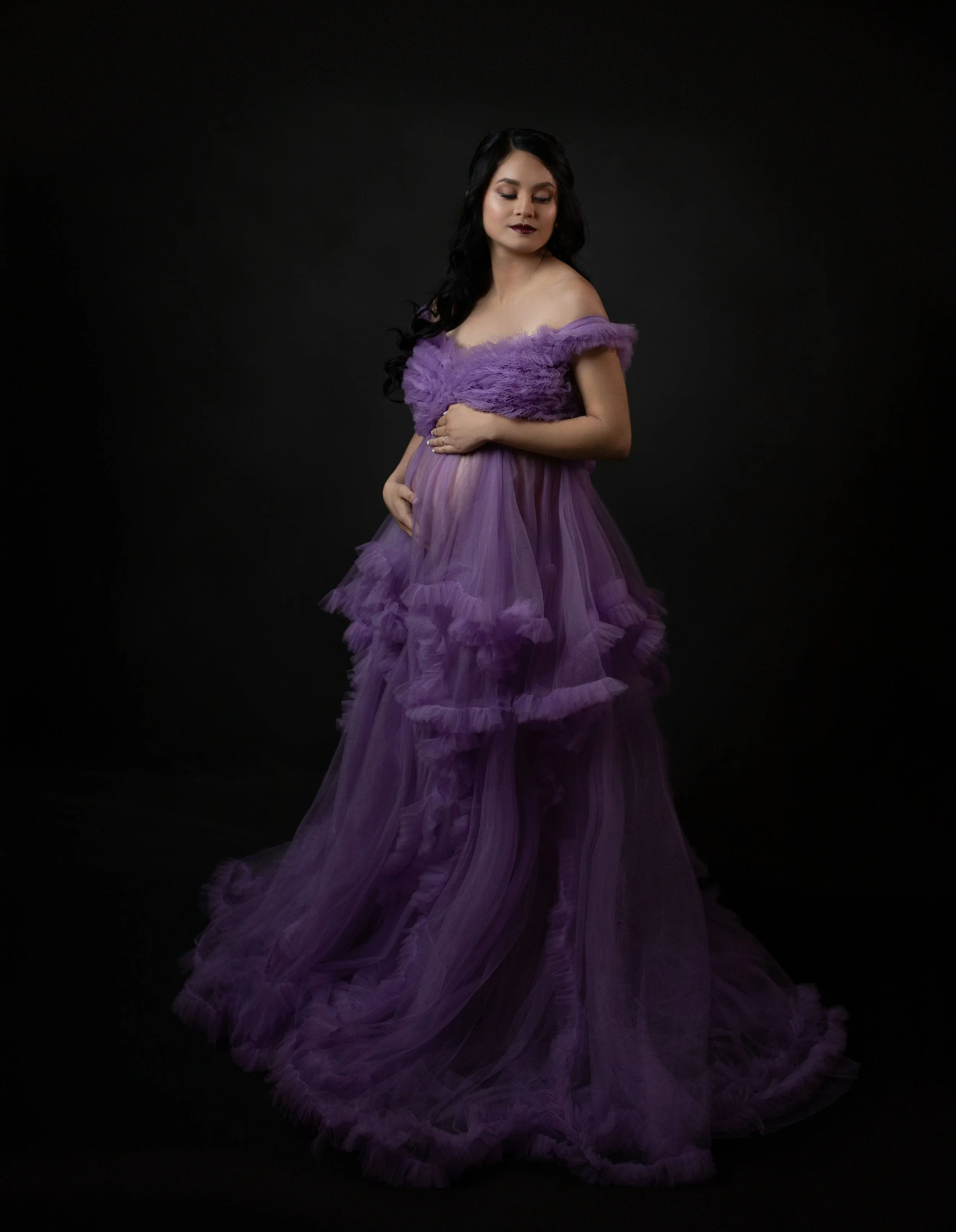 a pregnant woman wearing a purple ruffled gown holding her belly with both hands looking down to her left