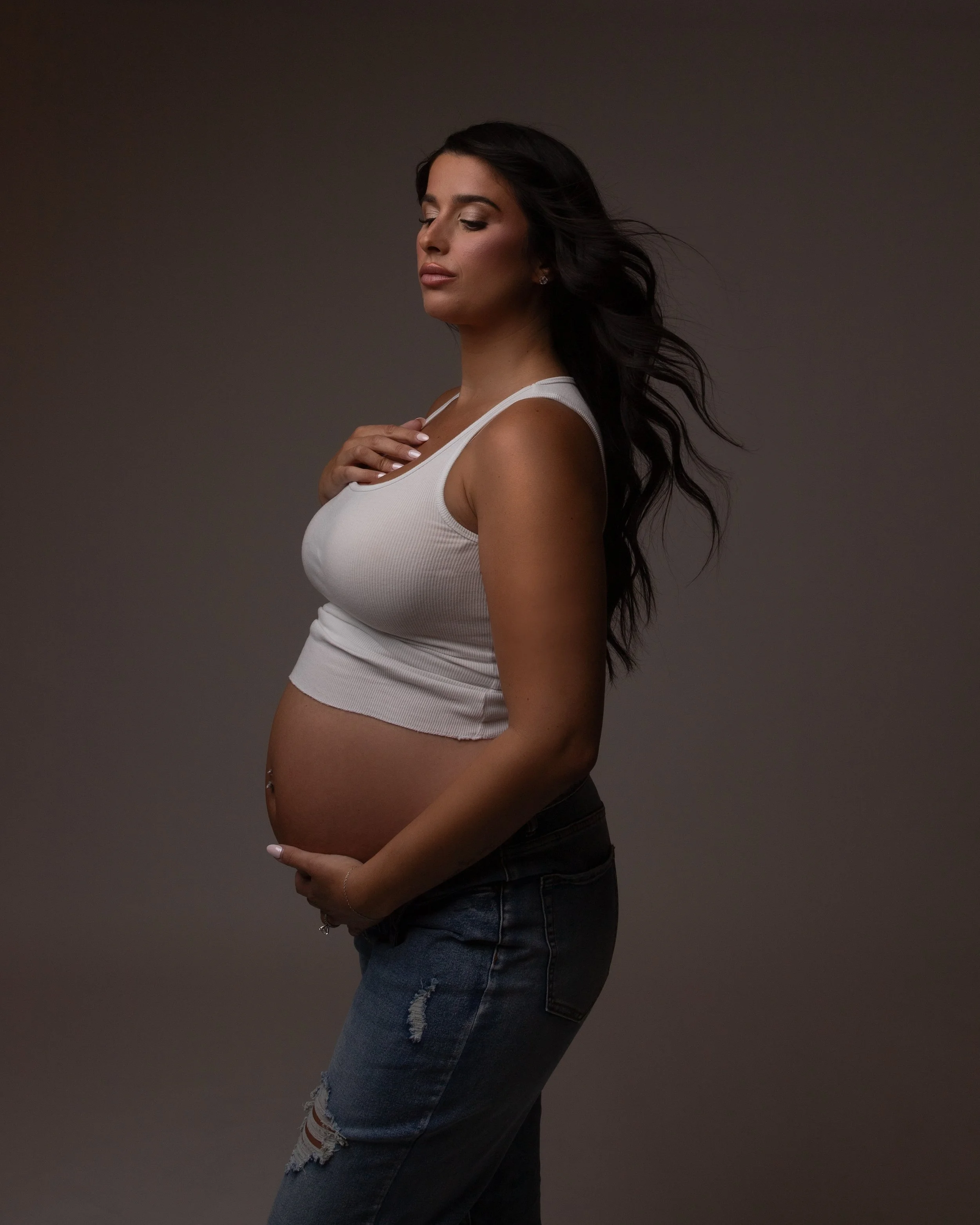 pregnant woman wearing a white tee shirt and jeans holidng her belly with her eyes closed and her hair blowing 