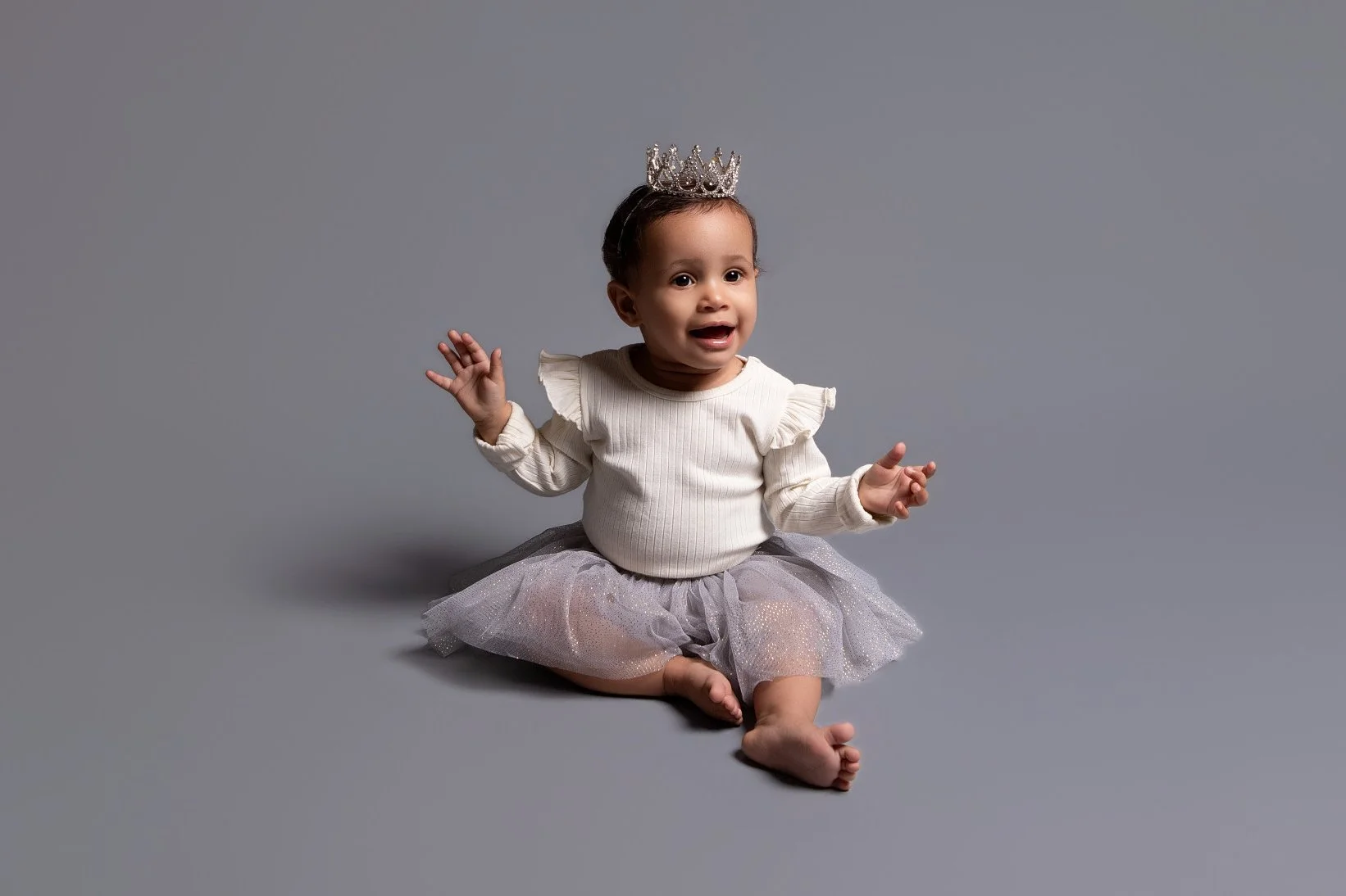 toddler girl wearing a cream colored onsie with grey background a silver tutu skirt hodling up both her hands and smiling