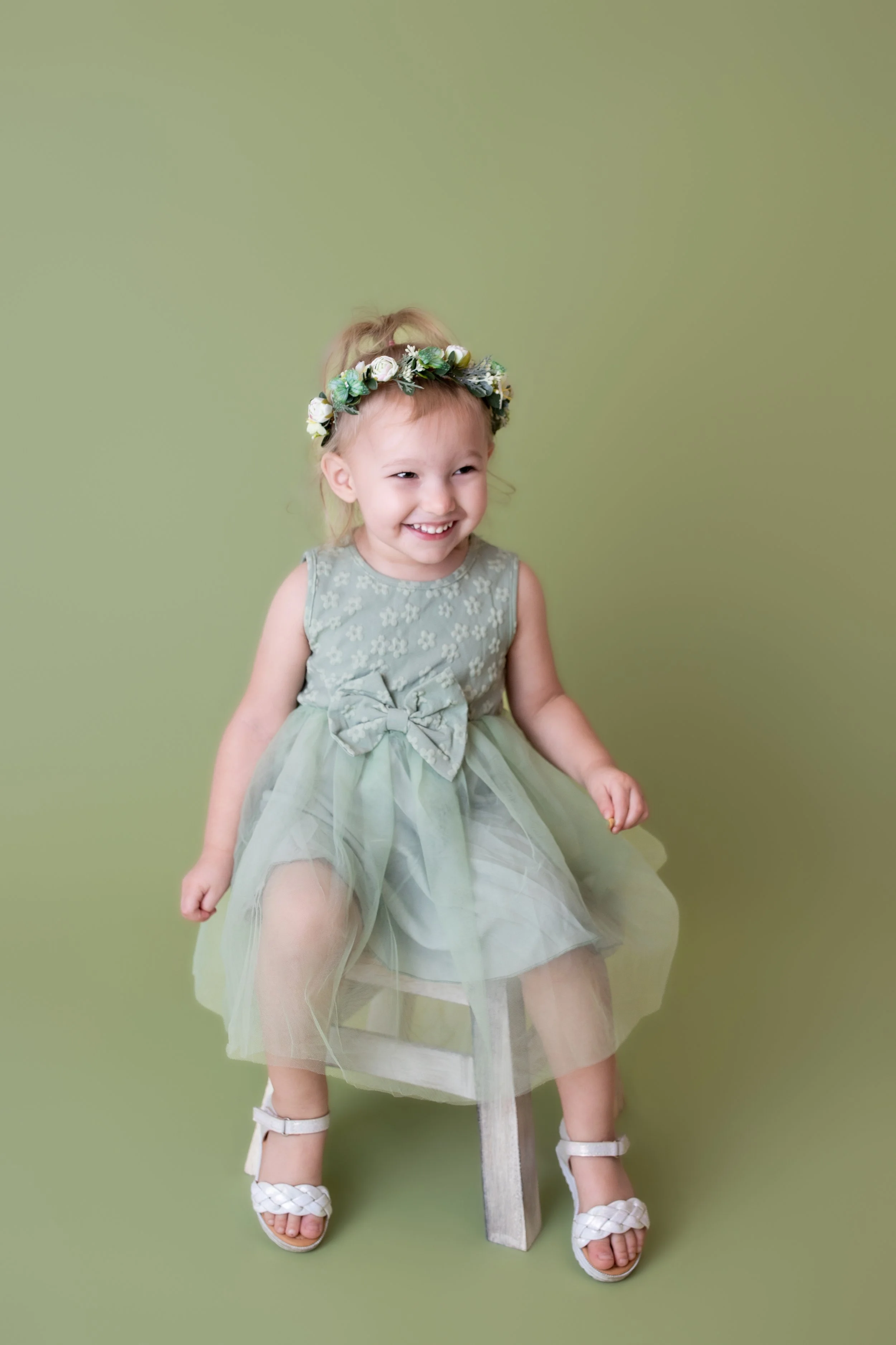 little girl with green floral wreathin her hair wearing a light green colored ruffled dress sitting on a light green colored box with a light green colored background