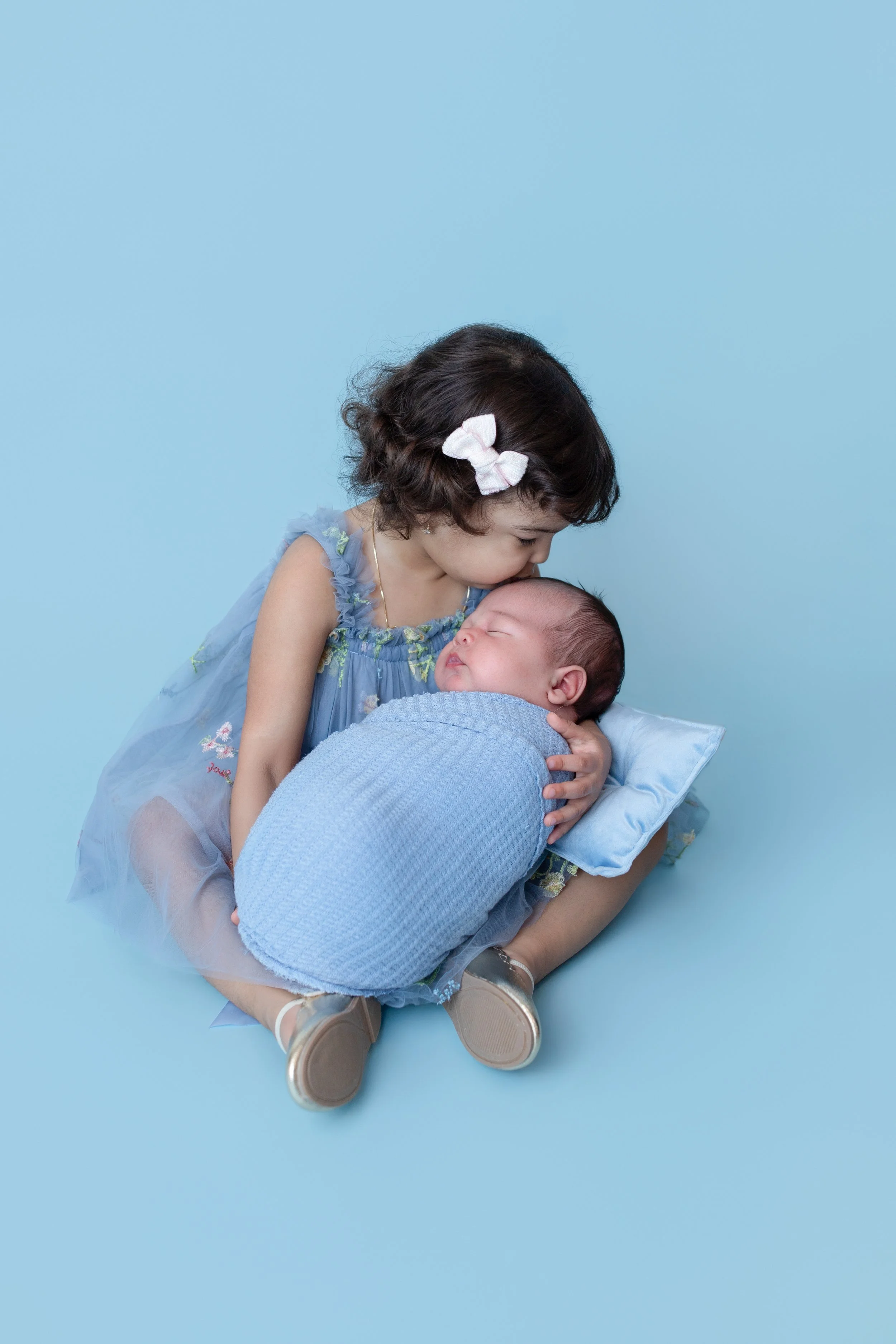 a two year old girl wearing a blue dress holidng  and kissing her newborn brother in a blue swaddle with a matching blue background 