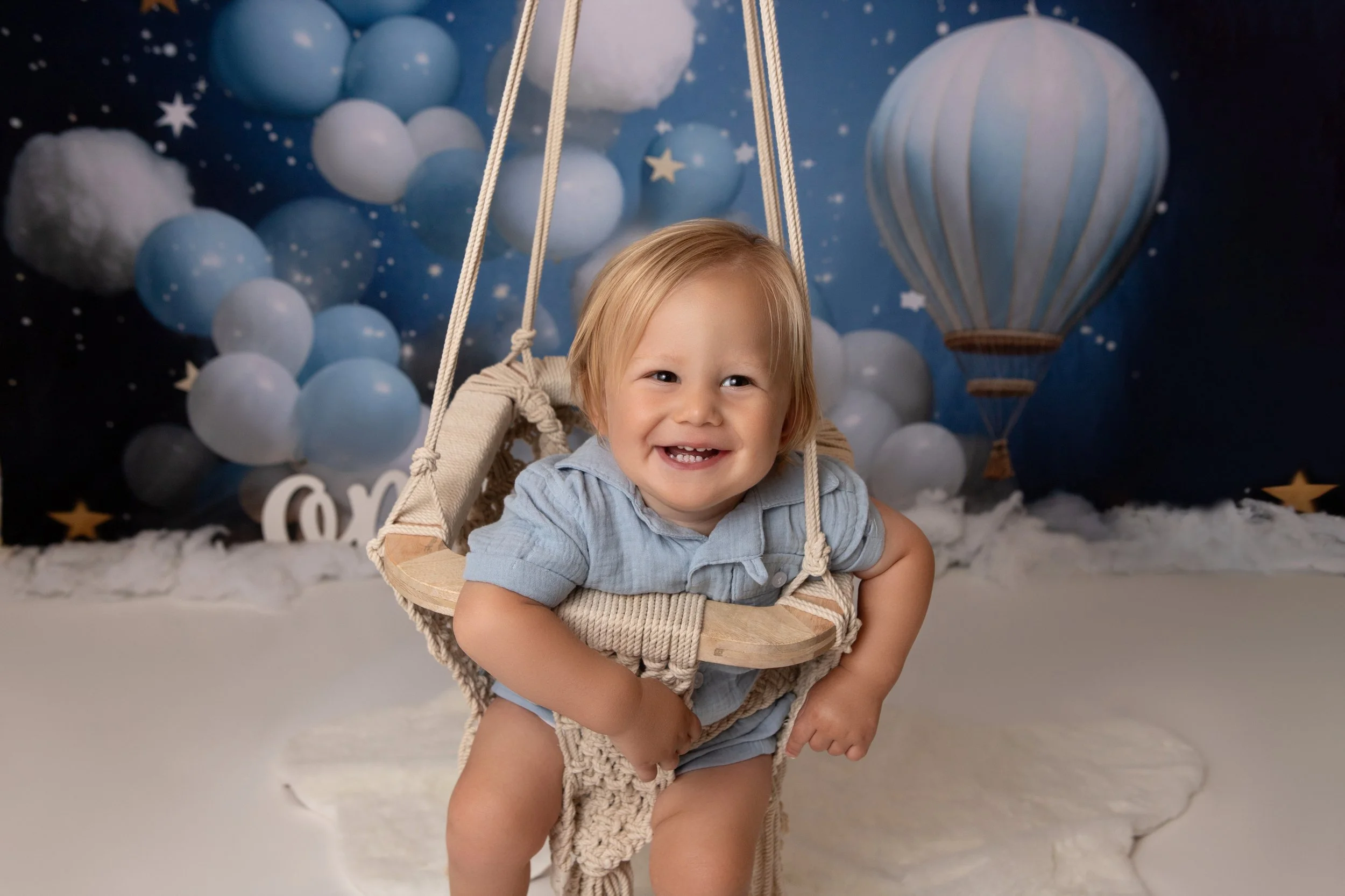 one year old boy in a swing with blue and white balloons in the background