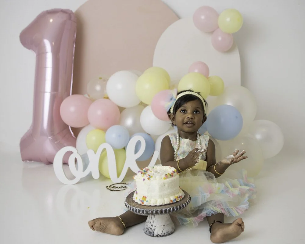 one year old girl sitting in front of a cake with colorful balloons 
