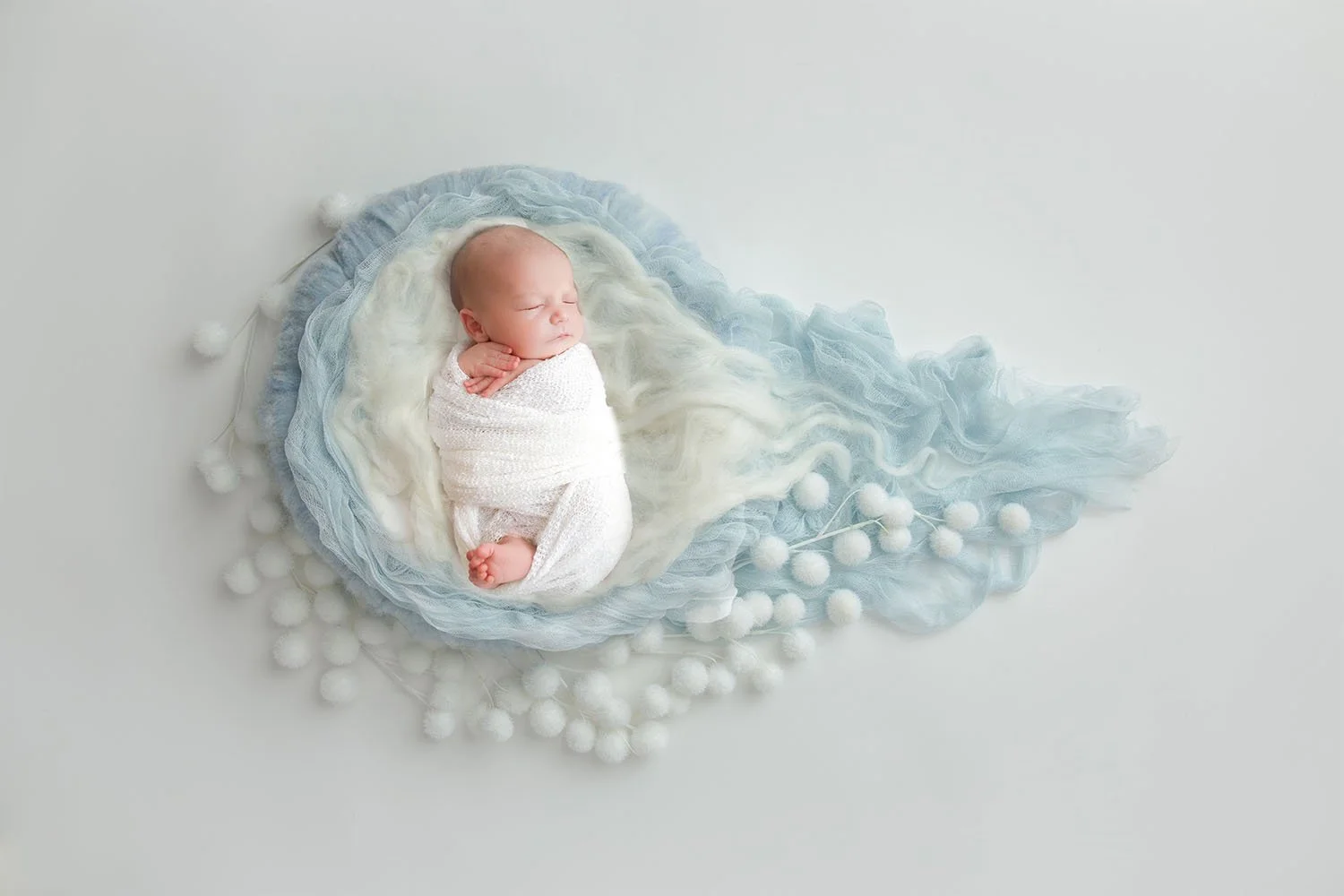 Sleeping newborn wrapped in a white swaddle, nestled in a blue and white cloud-like fabric with delicate pom-pom details. Dreamy and artistic newborn photography in Lancaster, PA.