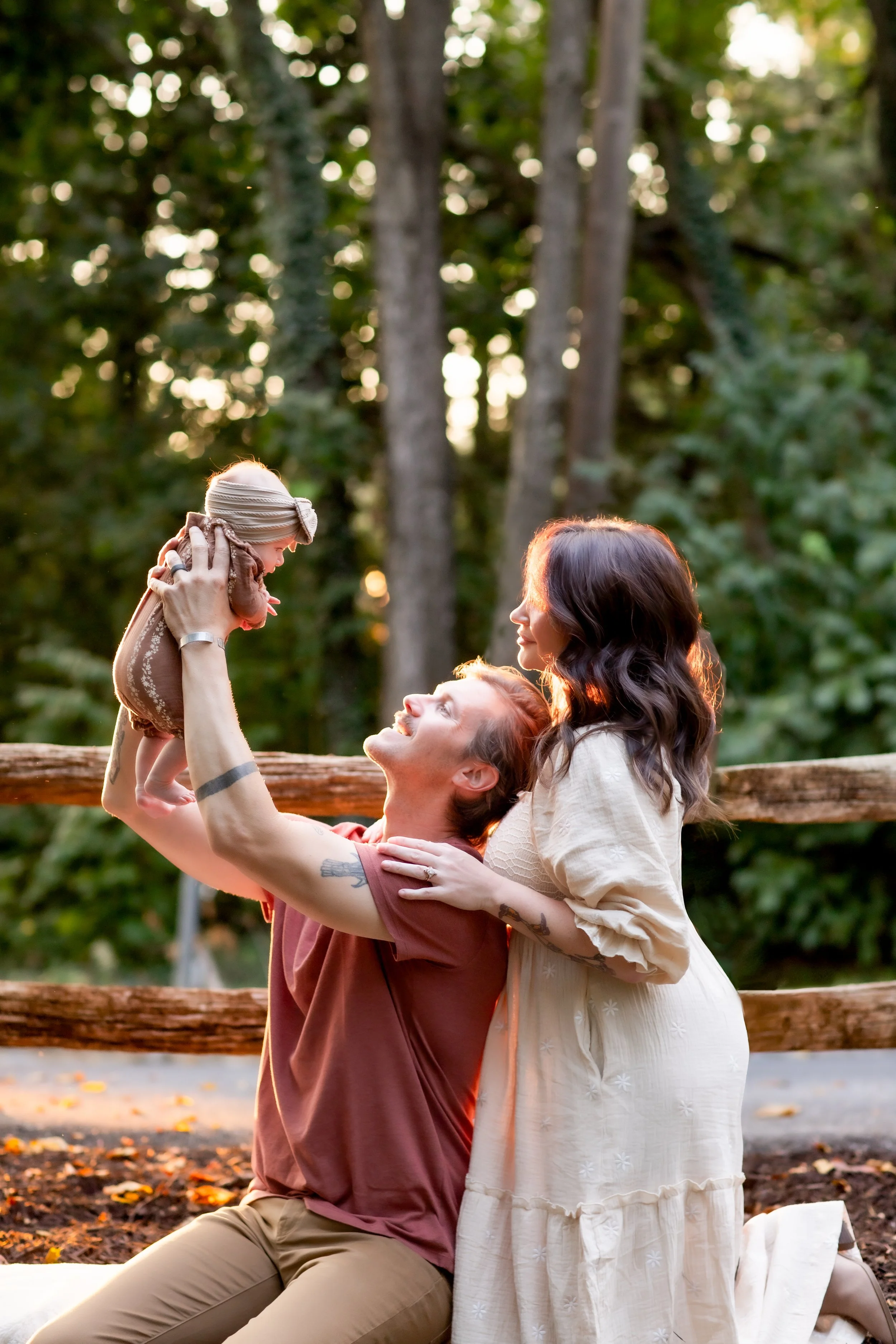 A family enjoying a moment outdoors in a forest during sunset, with a man holding a baby girl up in the air while two women watch and smile.