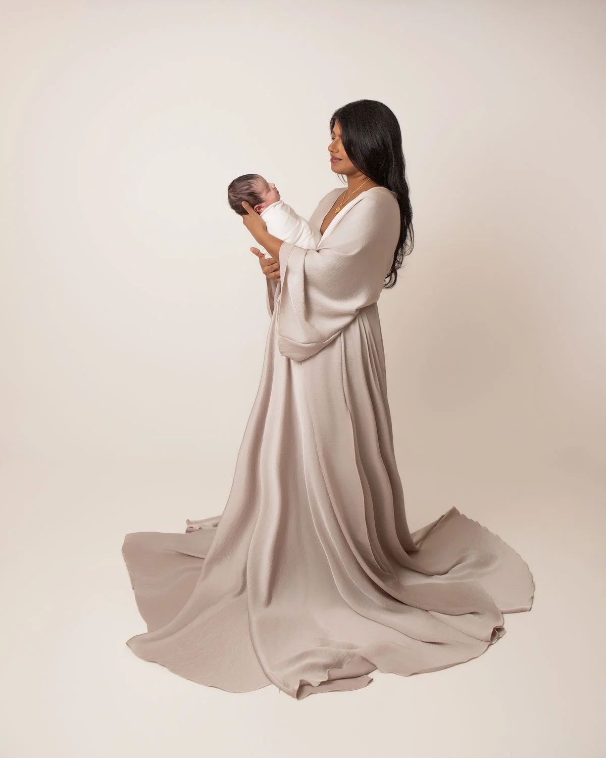 Mother in a flowing cream gown looking lovingly at her newborn baby, holding them close. Graceful and emotional motherhood photography in Lancaster, PA.