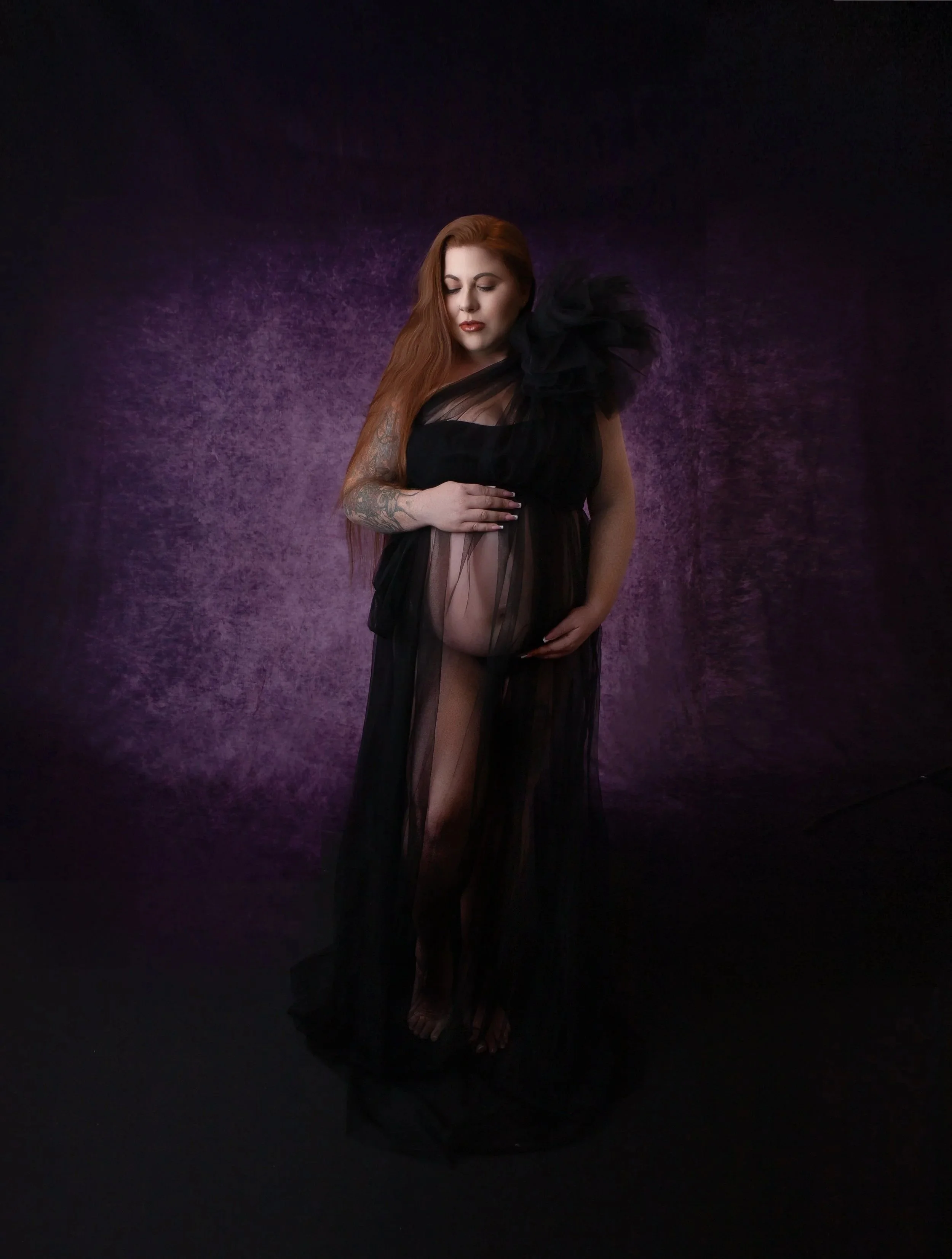 pregnant woman holding her belly wearing a black sheer gown with a purple background