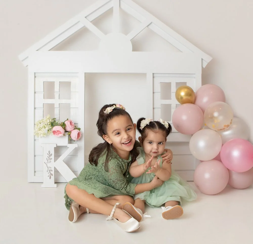 two littel girls smiling wearing green dresses with a white house and colorful balloons in the background