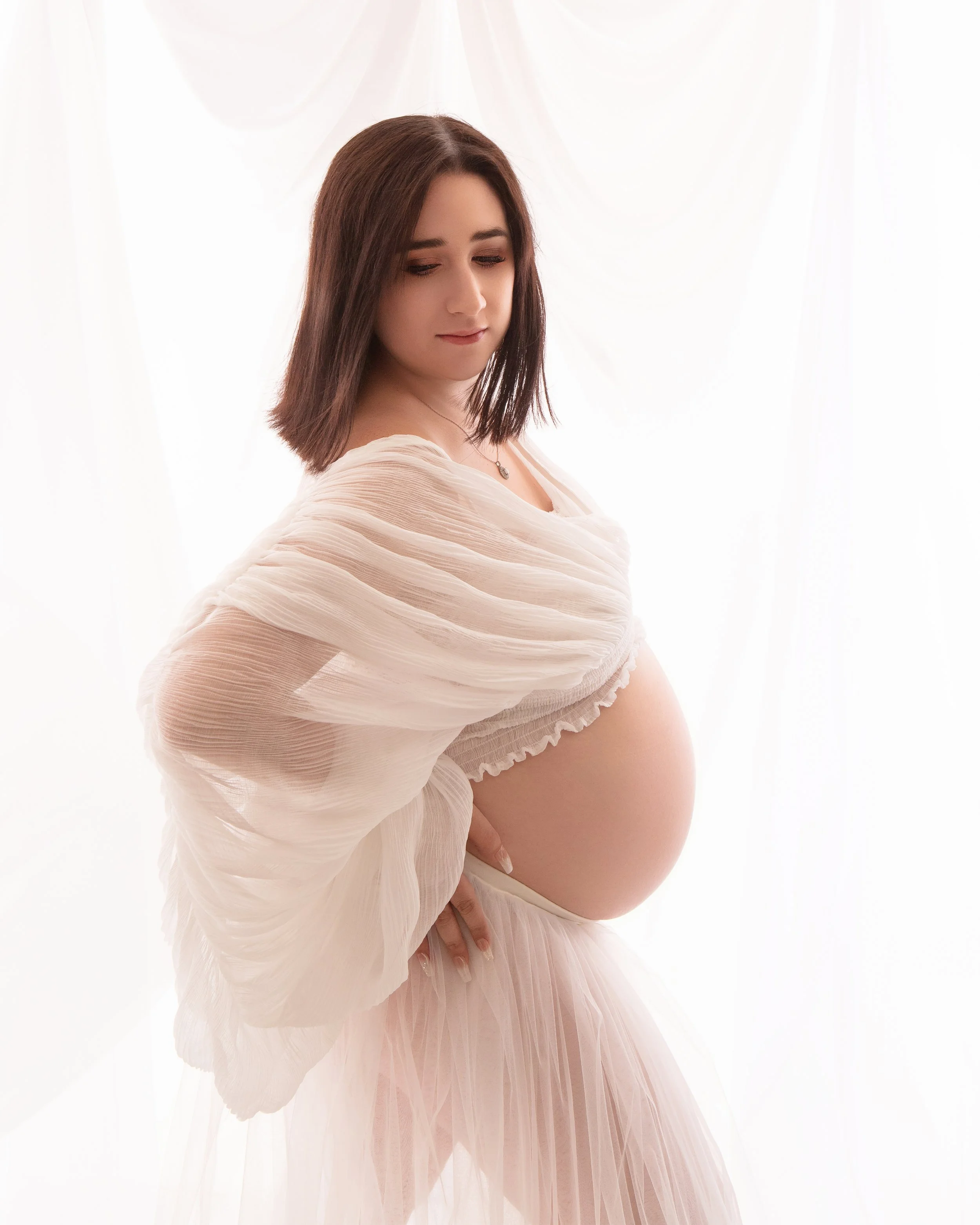 a pregnant women in a white sheet top with her hand on her hip looking down