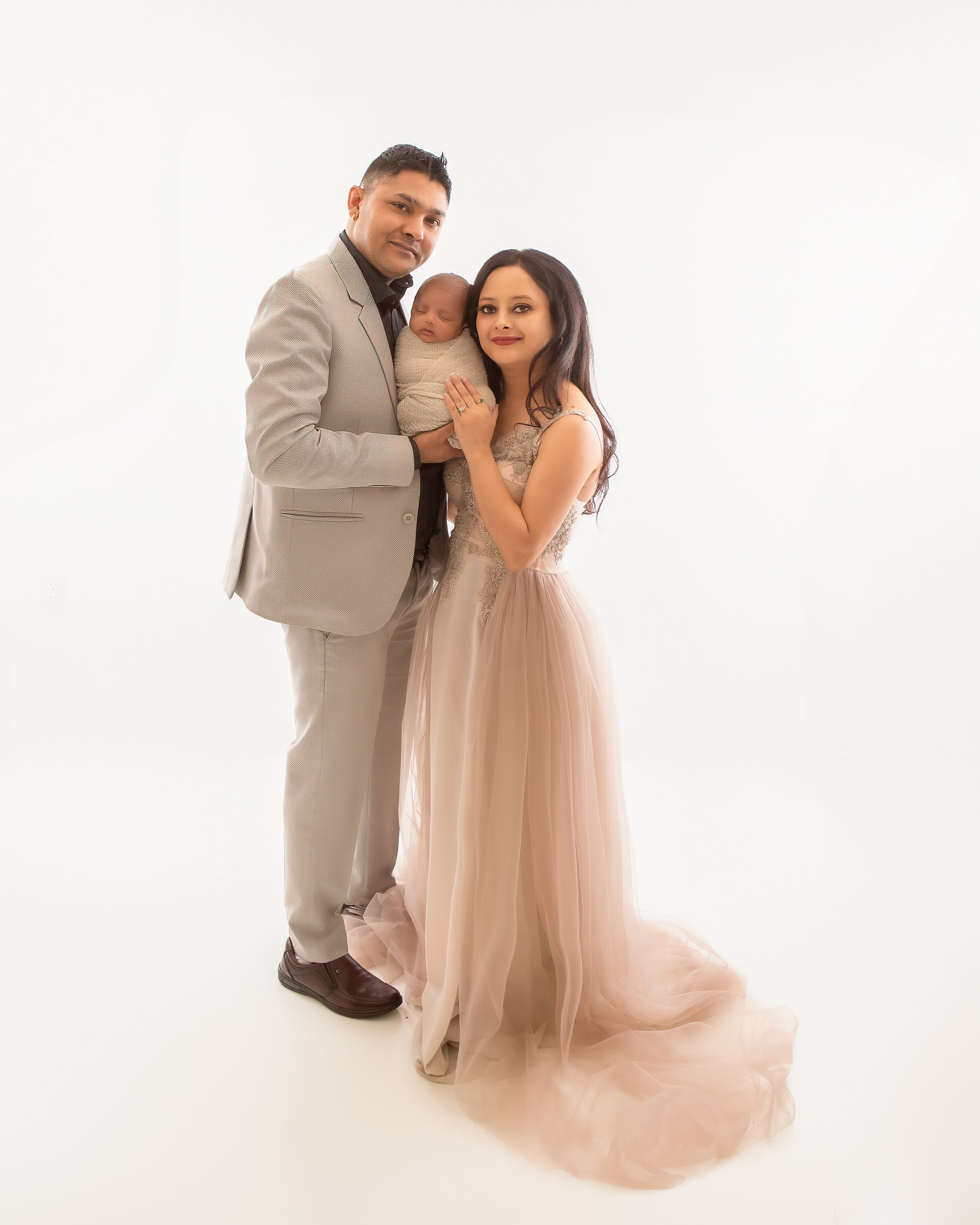 A family portrait of a man, woman, and baby against a plain white background. The man is wearing a light gray suit with a black shirt, the woman is wearing a beige dress with lace details, and the baby is wrapped in a beige blanket.